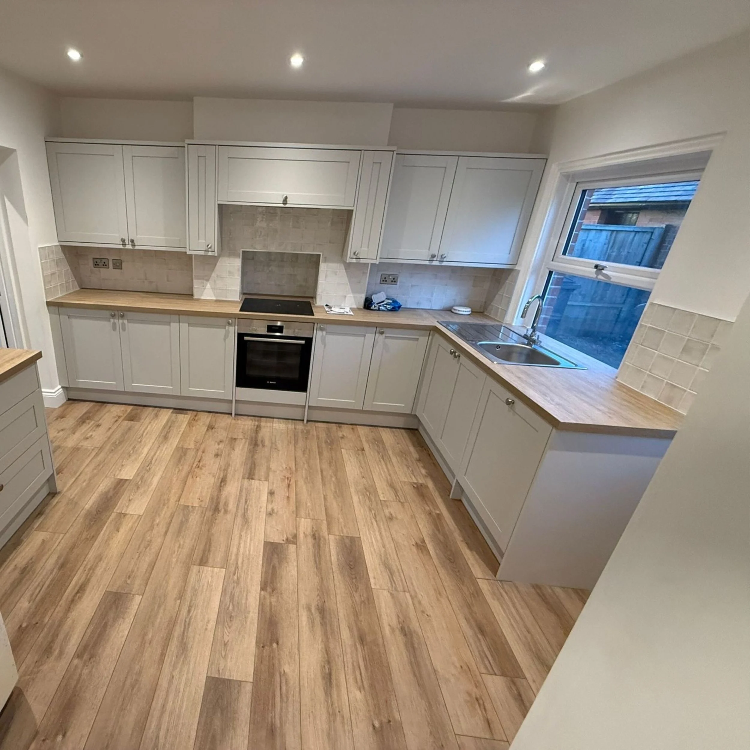 Kitchen with white cabinets, wooden countertops, a built-in oven, cooktop, stainless steel sink, window overlooking an outdoor area, beige tile backsplash, and wooden flooring.