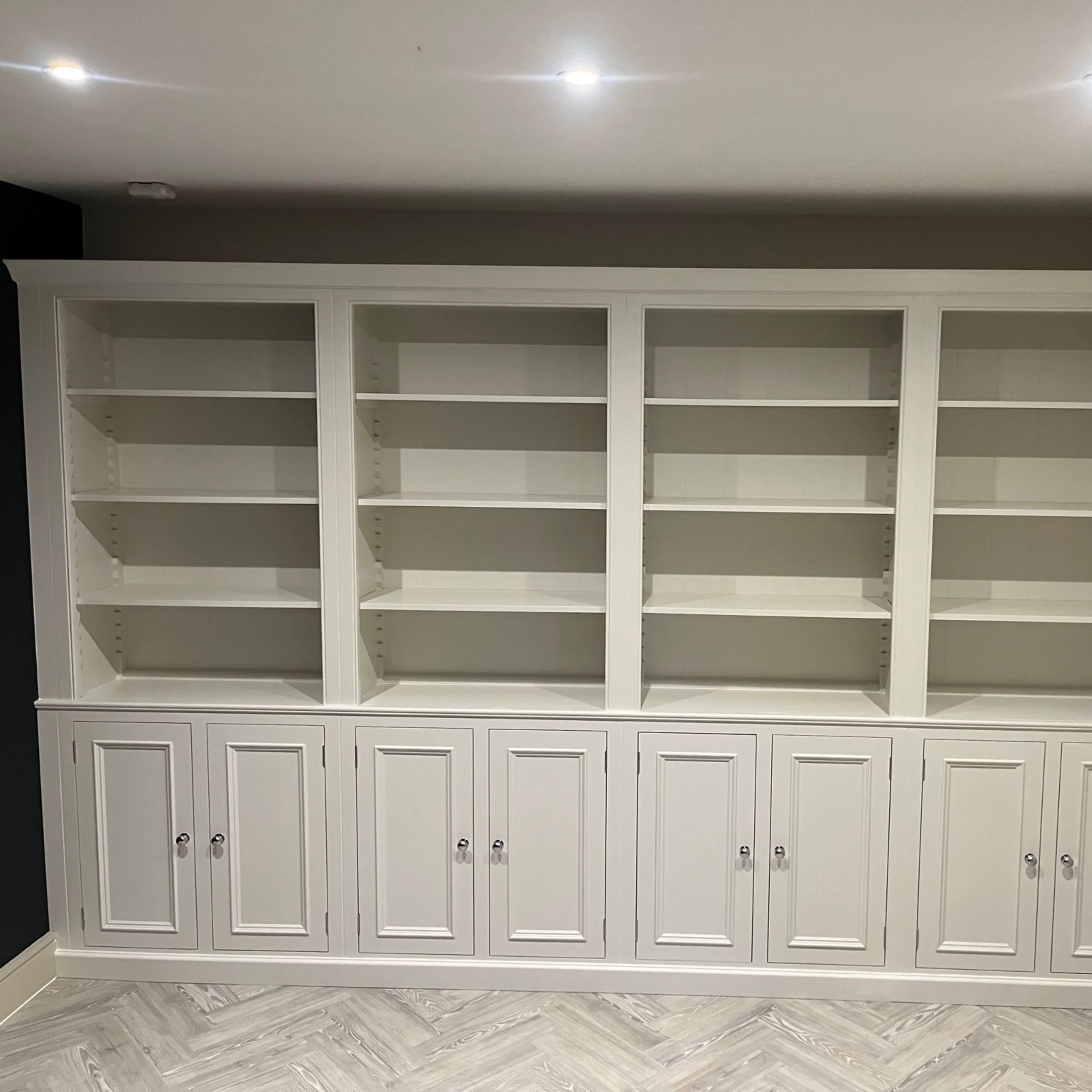 Empty white built-in bookshelf with closed cabinet doors at the bottom in a room with light-colored herringbone-patterned floor.