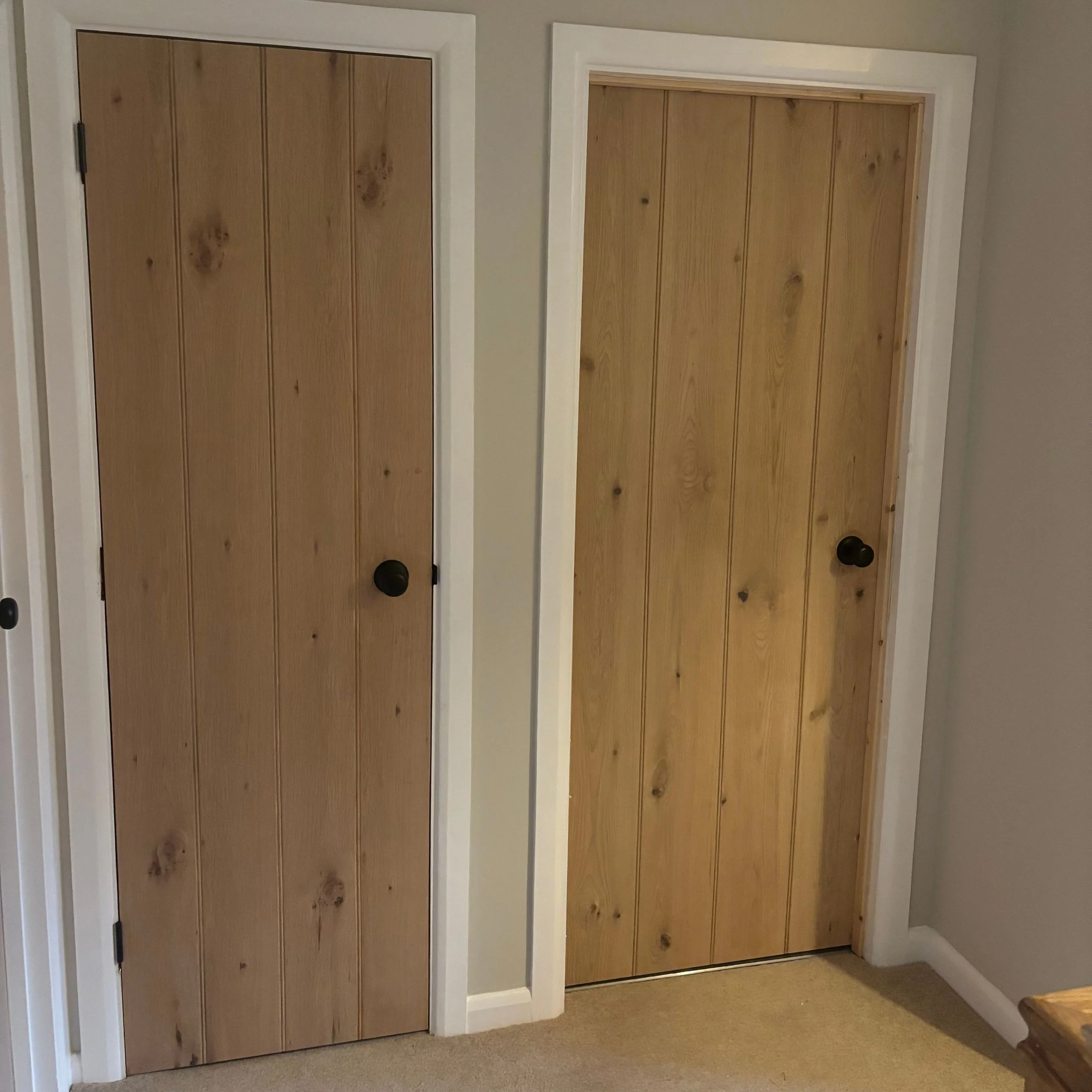 Two closed wooden doors, one on the left and one on the right, with white trim and black doorknobs, in a beige-carpeted room.
