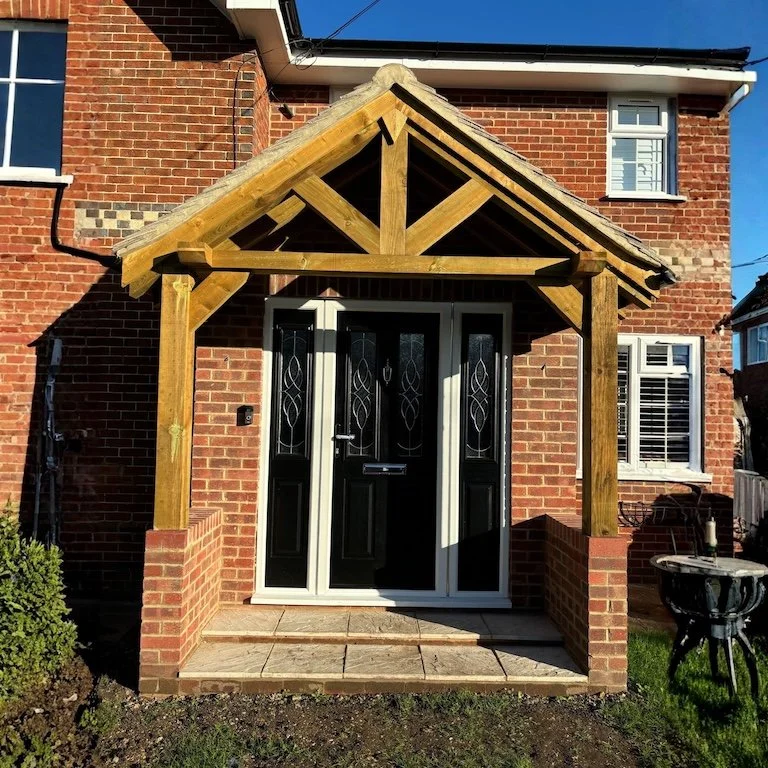 New wooden porch frame with brick pillars, installed in front of a house with a black door and glass panels, with a small tiled step and some ground work in progress.