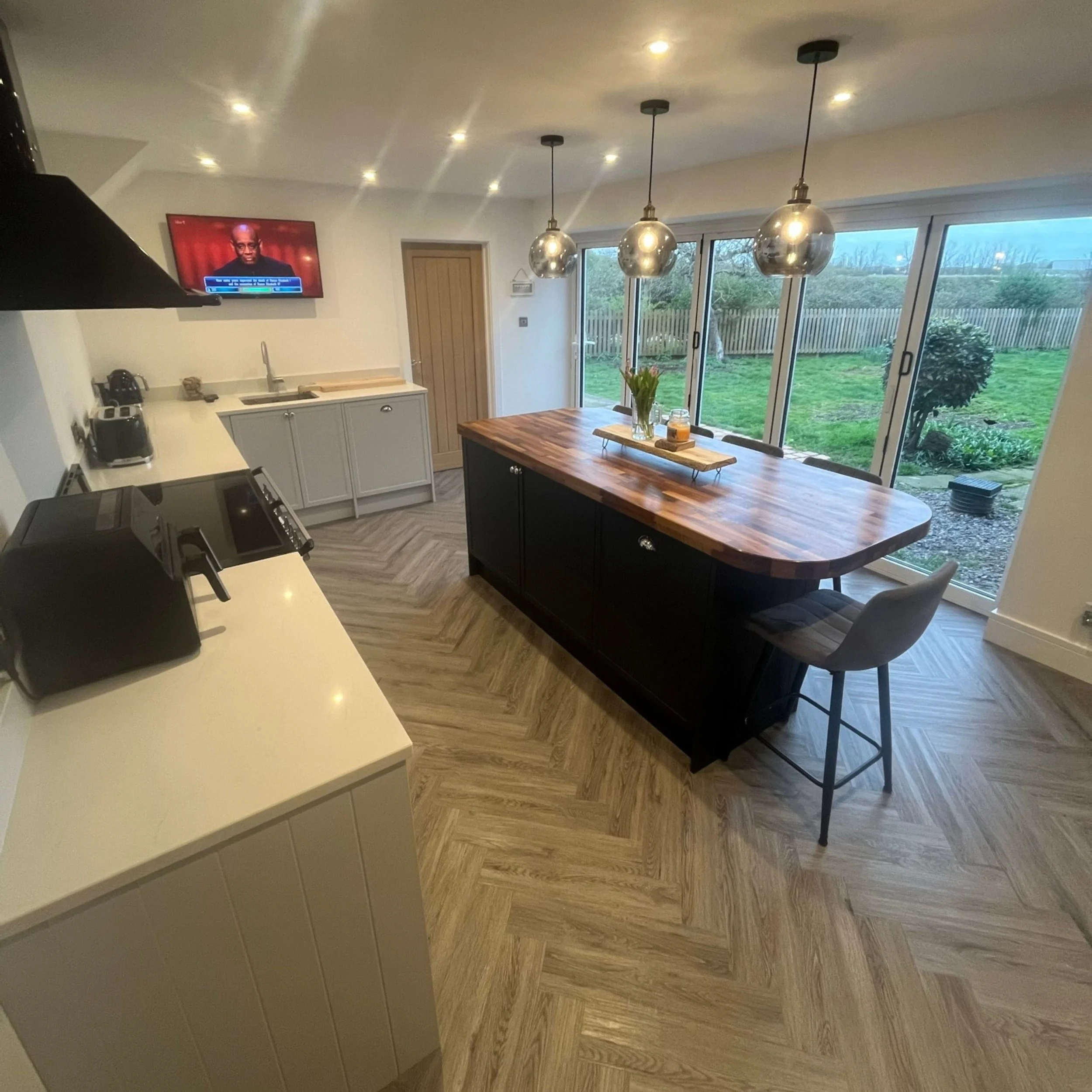 A modern kitchen with a central island, pendant lights, large sliding glass doors overlooking a backyard, a wall-mounted TV, and various kitchen appliances.
