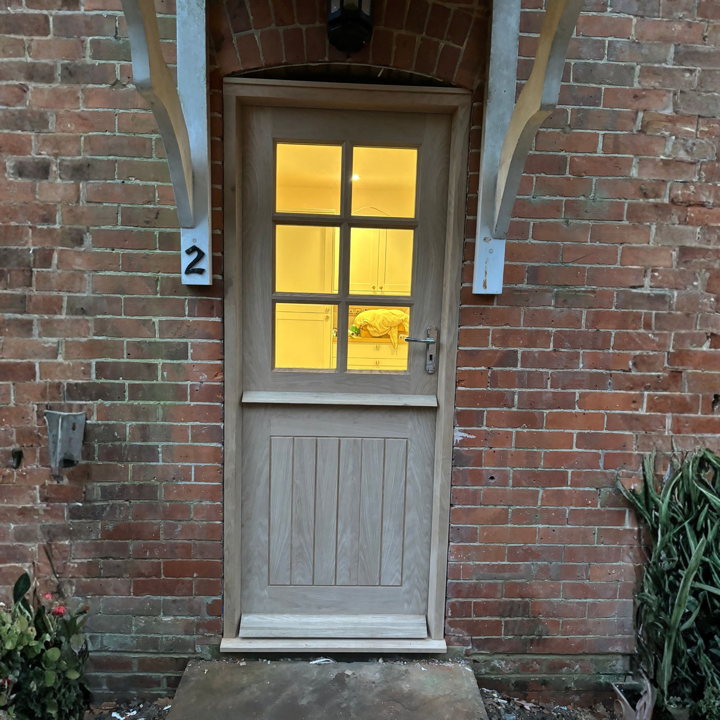 Wooden door with glass panes, illuminated by warm yellow light from inside, set in a brick wall with metal brackets and house number 2.