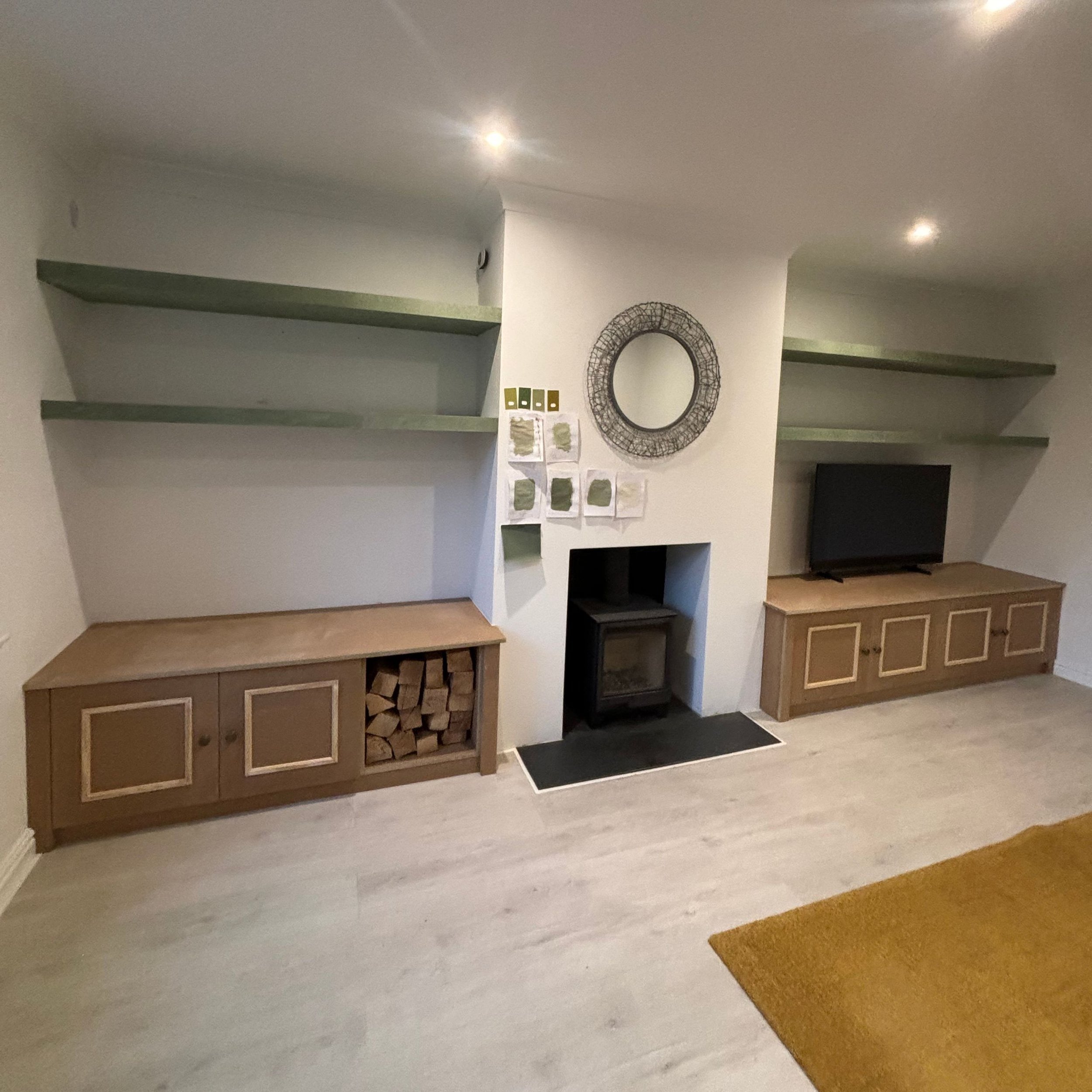 Living room with two wooden cabinets beneath open green shelves, a small fireplace in the center, a round mirror above, and a flat-screen TV on the right cabinet. Wall decor includes art pieces and a circular wireframe mirror.