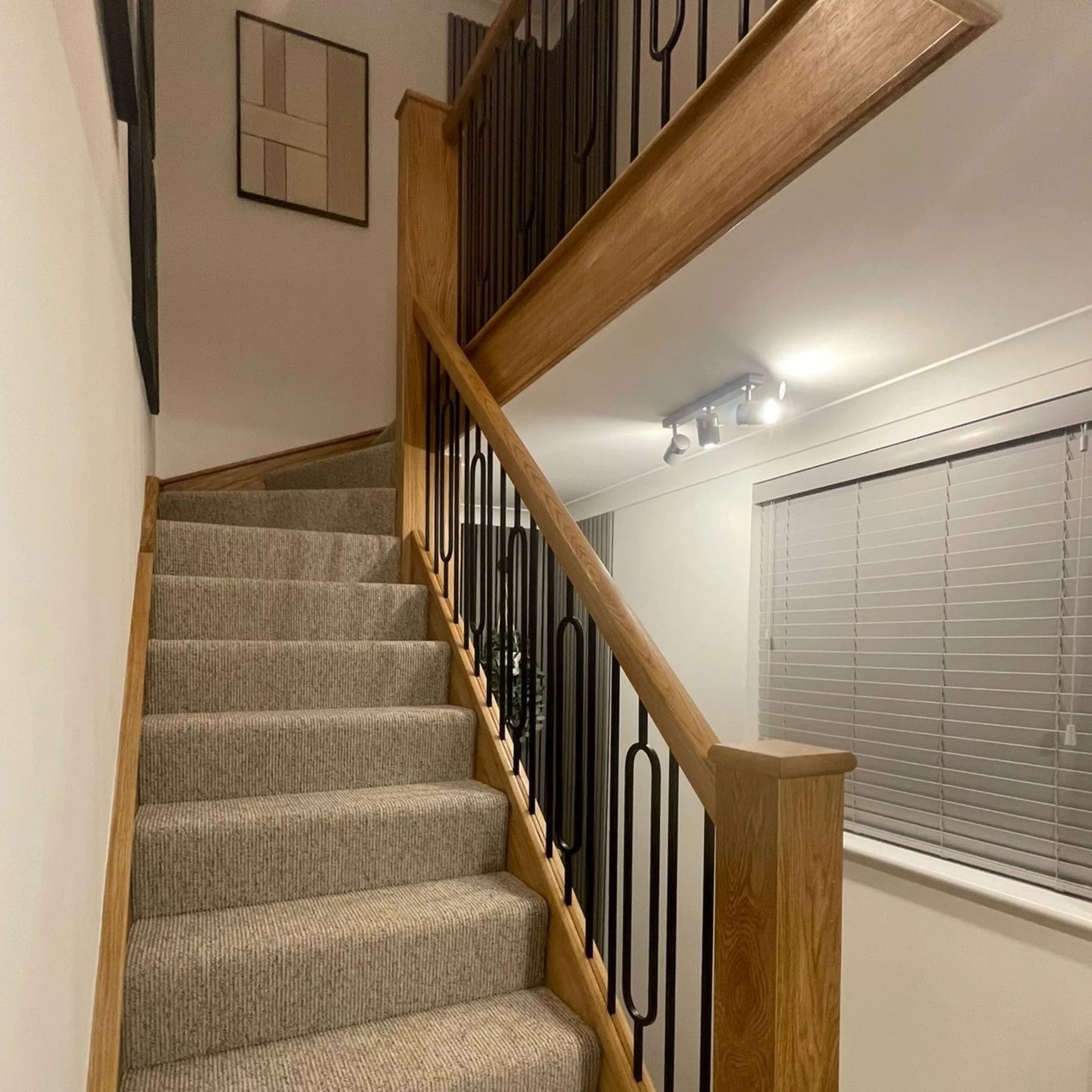 Indoor staircase with carpeted steps, wooden handrail, and black metal balusters leading to an upper level. A window with closed blinds on the right and a wall-mounted light fixture overhead.