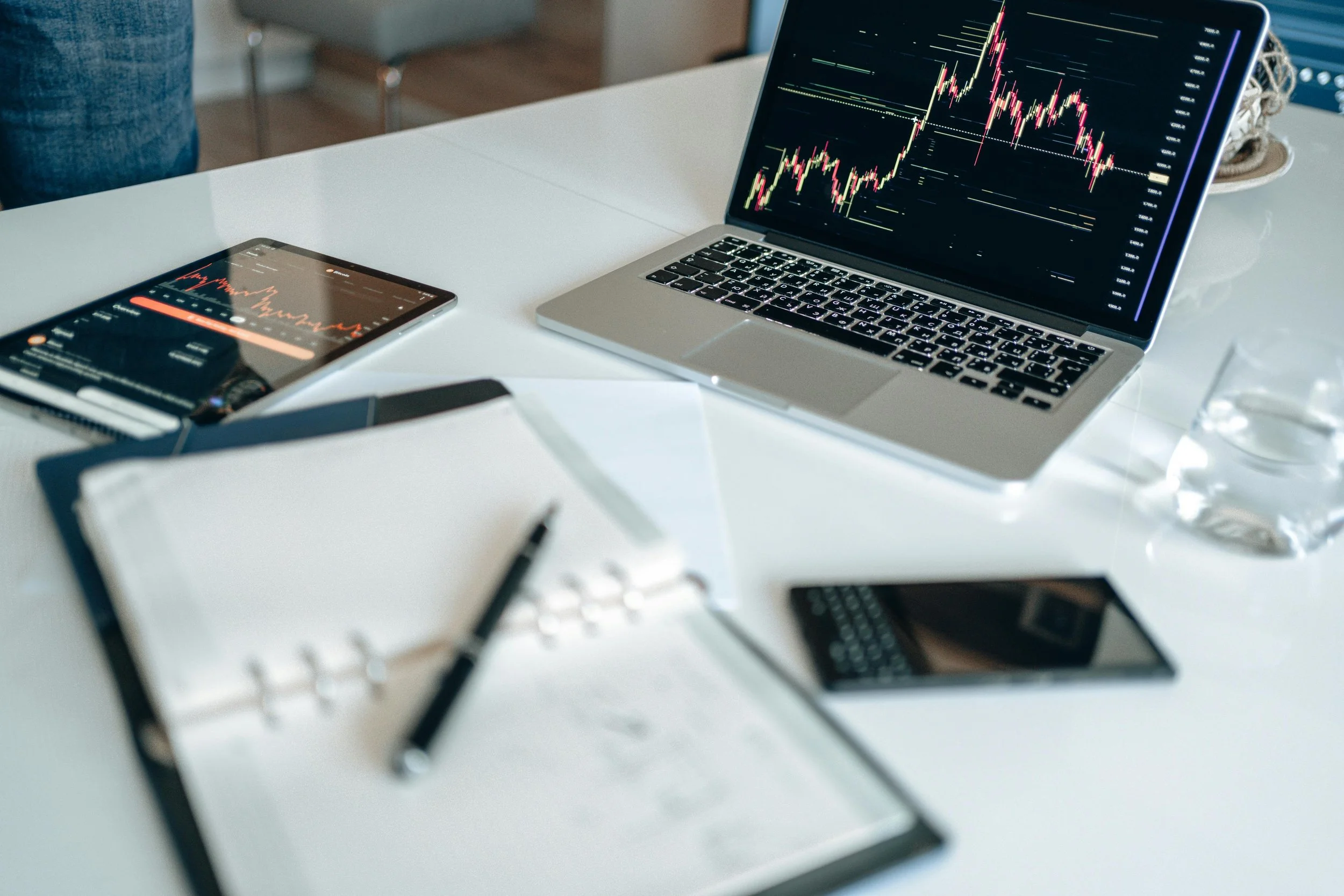 Laptop and tablet on a desk displaying stock market charts, with notebooks, pen, smartphone, and glass of water nearby.