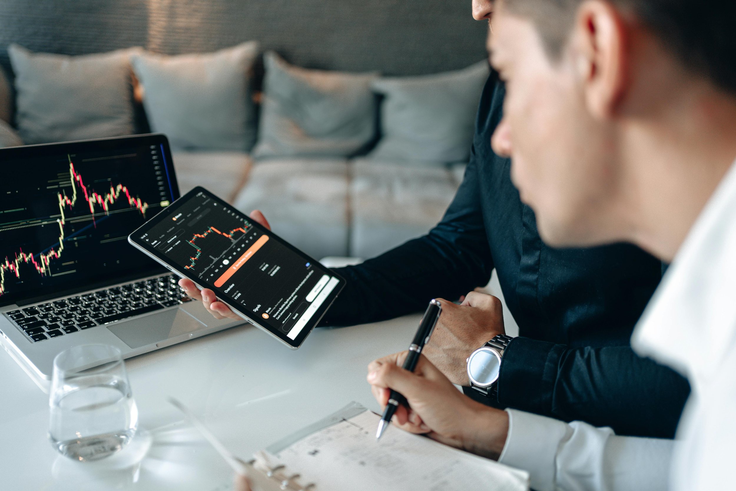 Two people reviewing stock market charts on a laptop, tablet, and notebook in a meeting or workspace setting, with a glass of water on the table.