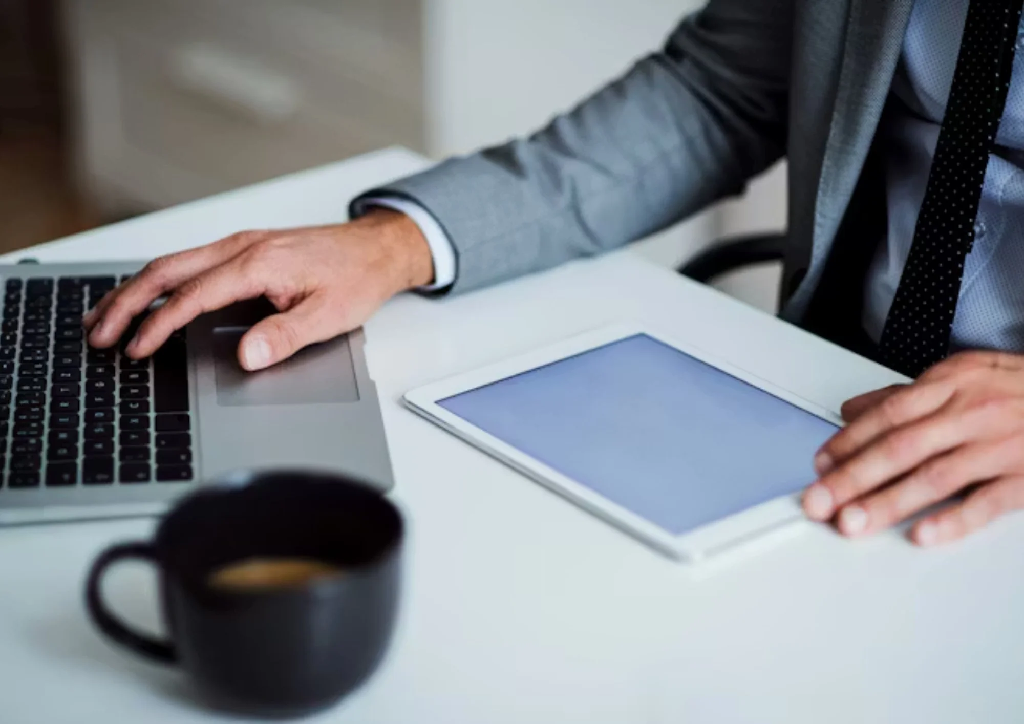 A person in a gray suit with a tie working at a white desk with a laptop, tablet, and a coffee cup.