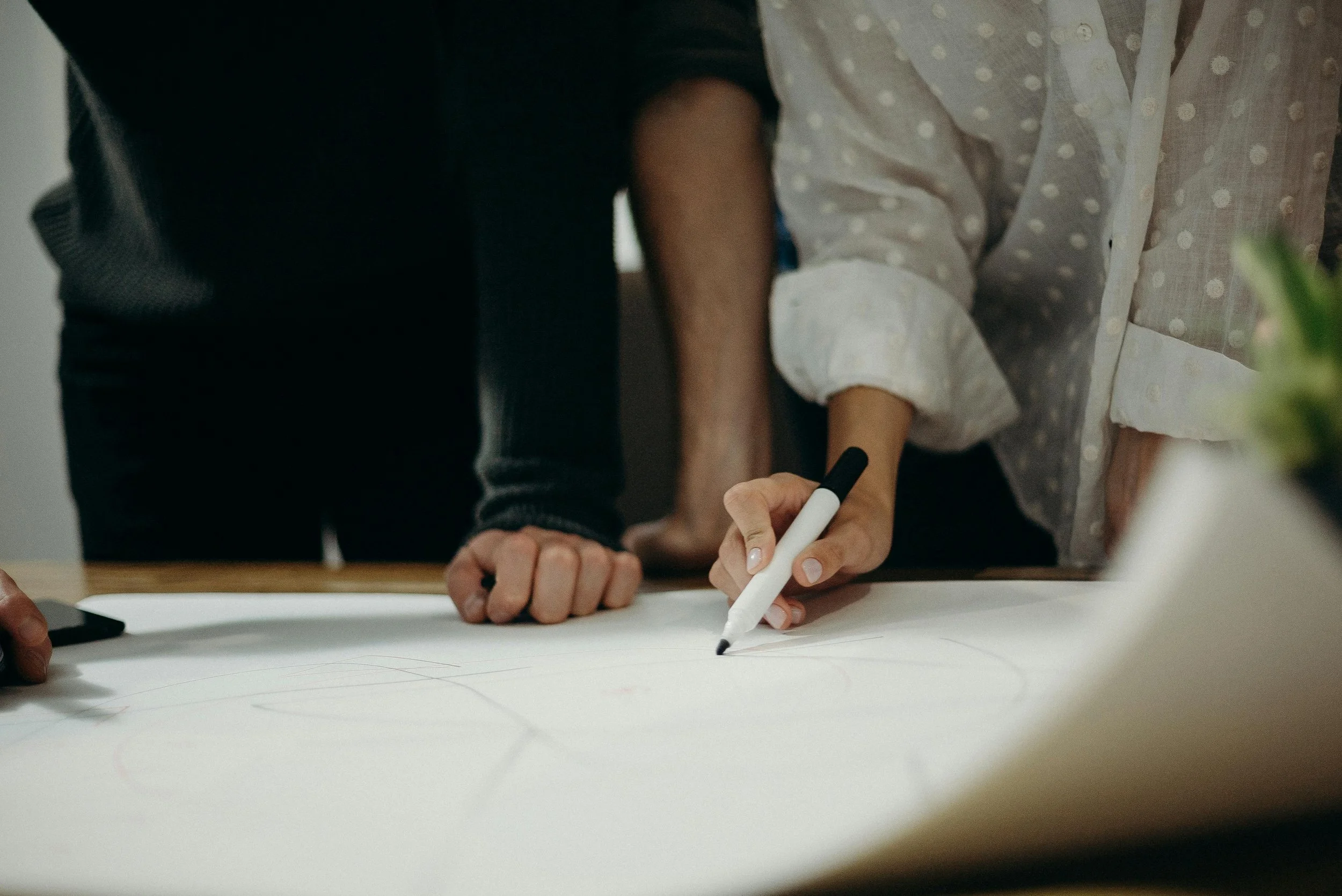 Two people are collaborating over a large sheet of paper. One person, wearing a long sleeve shirt with a floral pattern, is writing or drawing with a white marker. The other person's hand is resting on the paper.