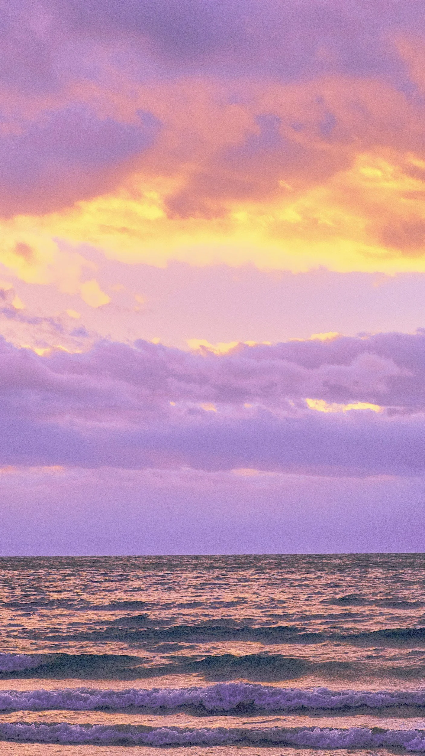 A sunset over the ocean with pink, purple, and yellow clouds in the sky and gentle waves on the water.
