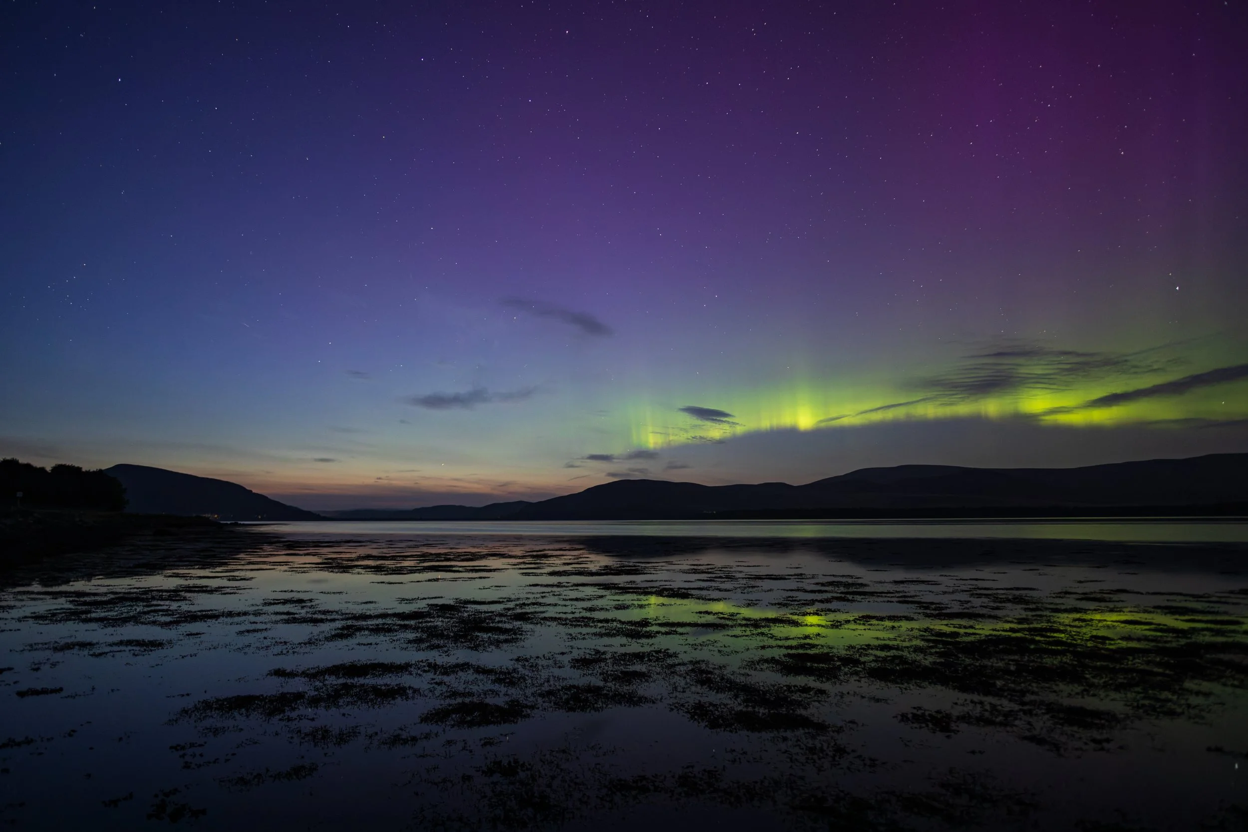 Nighttime landscape with the northern lights over a lake, mountains in the distance, and a starry sky.