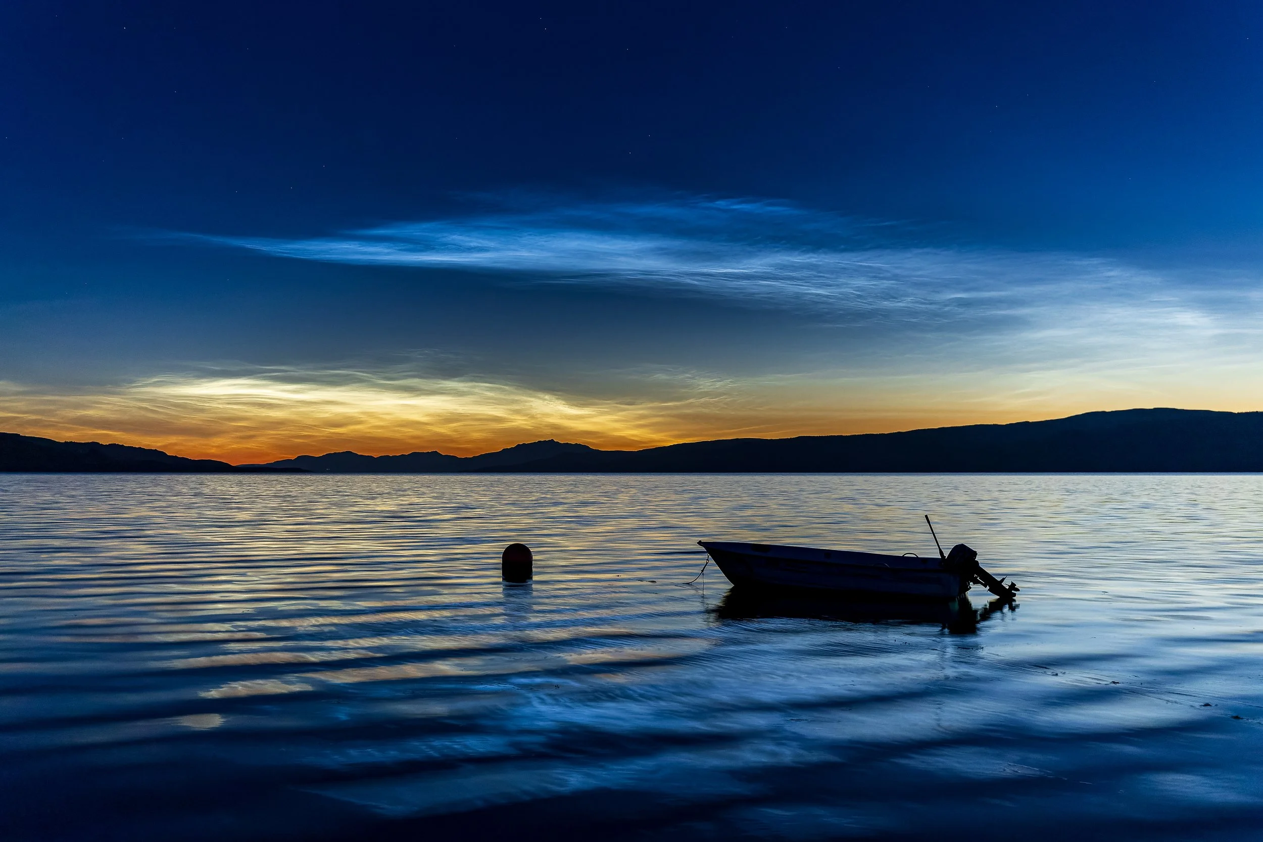 A small boat floats on calm water at sunset, with a buoy nearby and mountains in the distance under a sky transitioning from warm orange to deep blue.