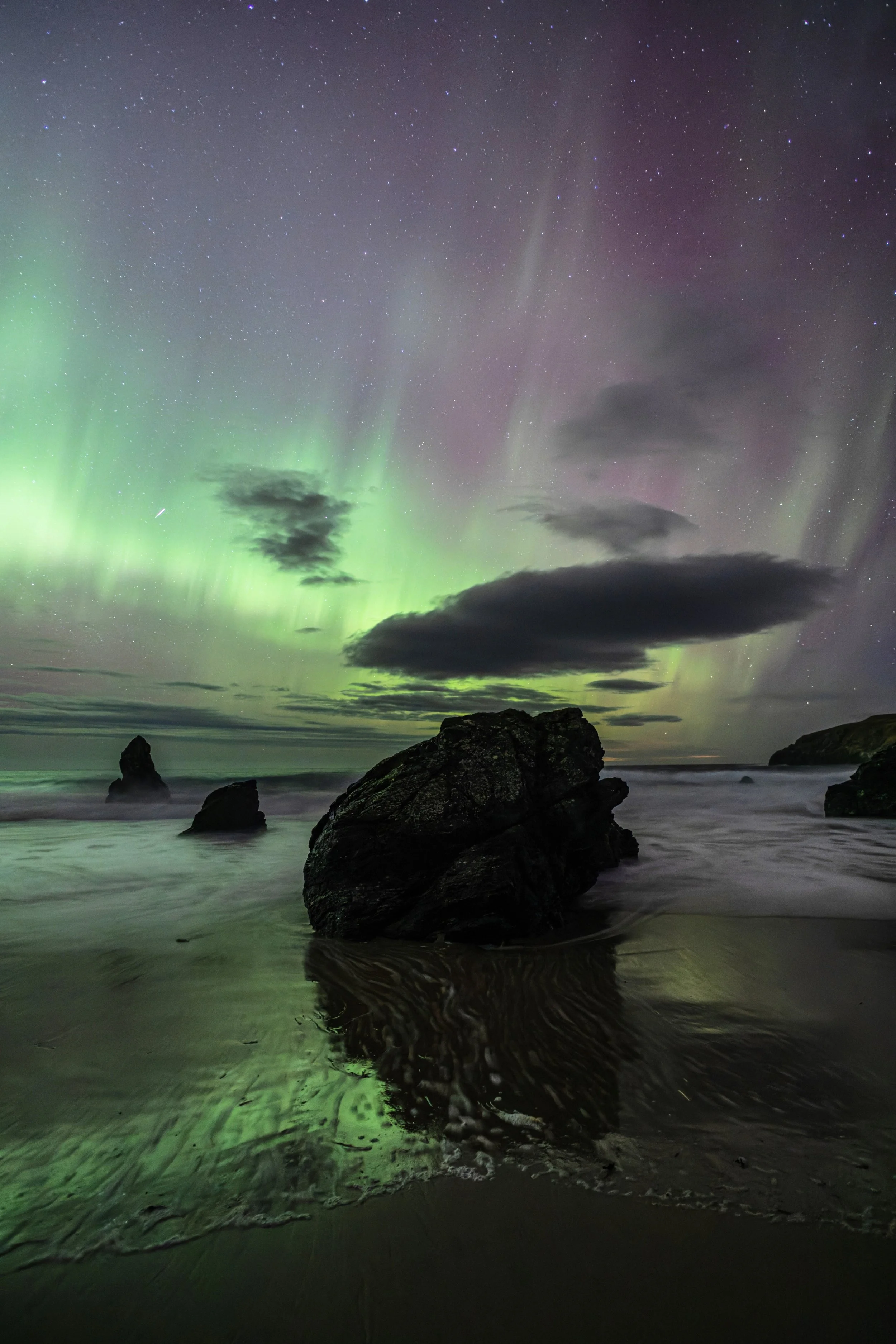 Nighttime coastal scene with large rocks on the beach and northern lights in the sky.