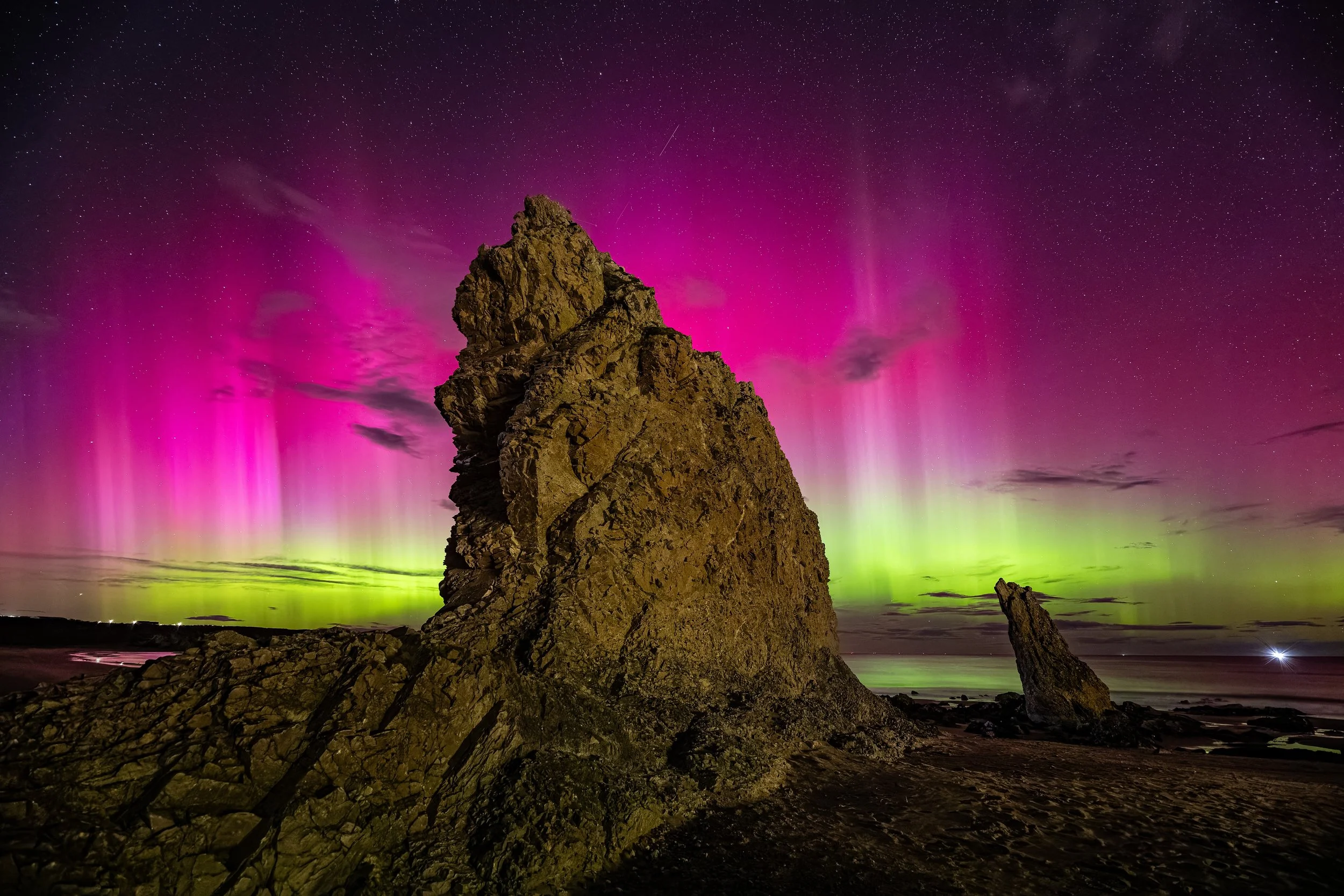 Nighttime scene at the beach featuring a large rock formation, with the sky illuminated by vibrant pink and green northern lights and visible stars.