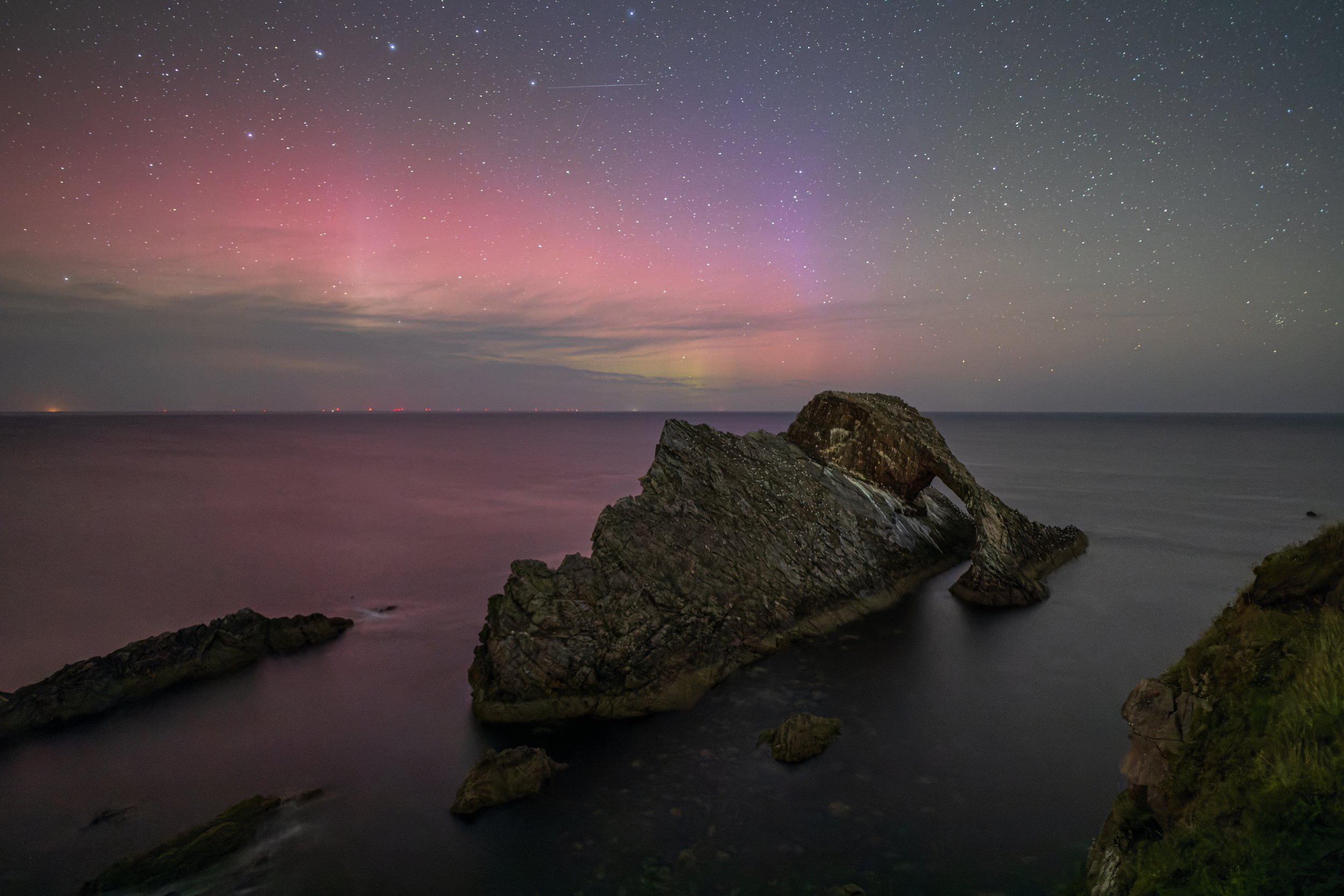 Nighttime coastal scene with large rock formations in the water, a colorful aurora borealis in the sky, and a star-filled night sky above.