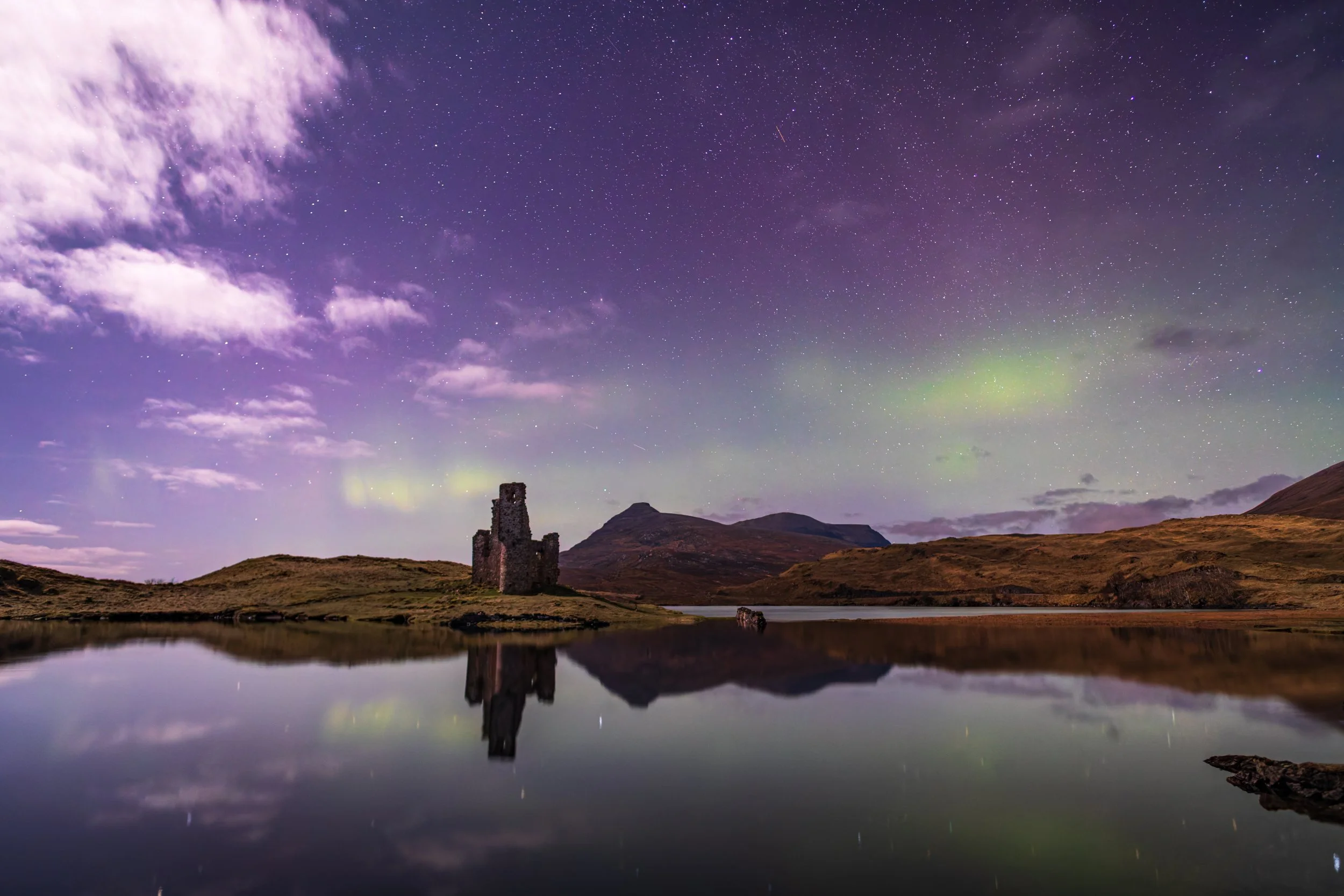 Nighttime landscape with starry sky, aurora borealis, an old stone castle ruin near a calm lake, mountains in the background, and reflection of the scene on the water.