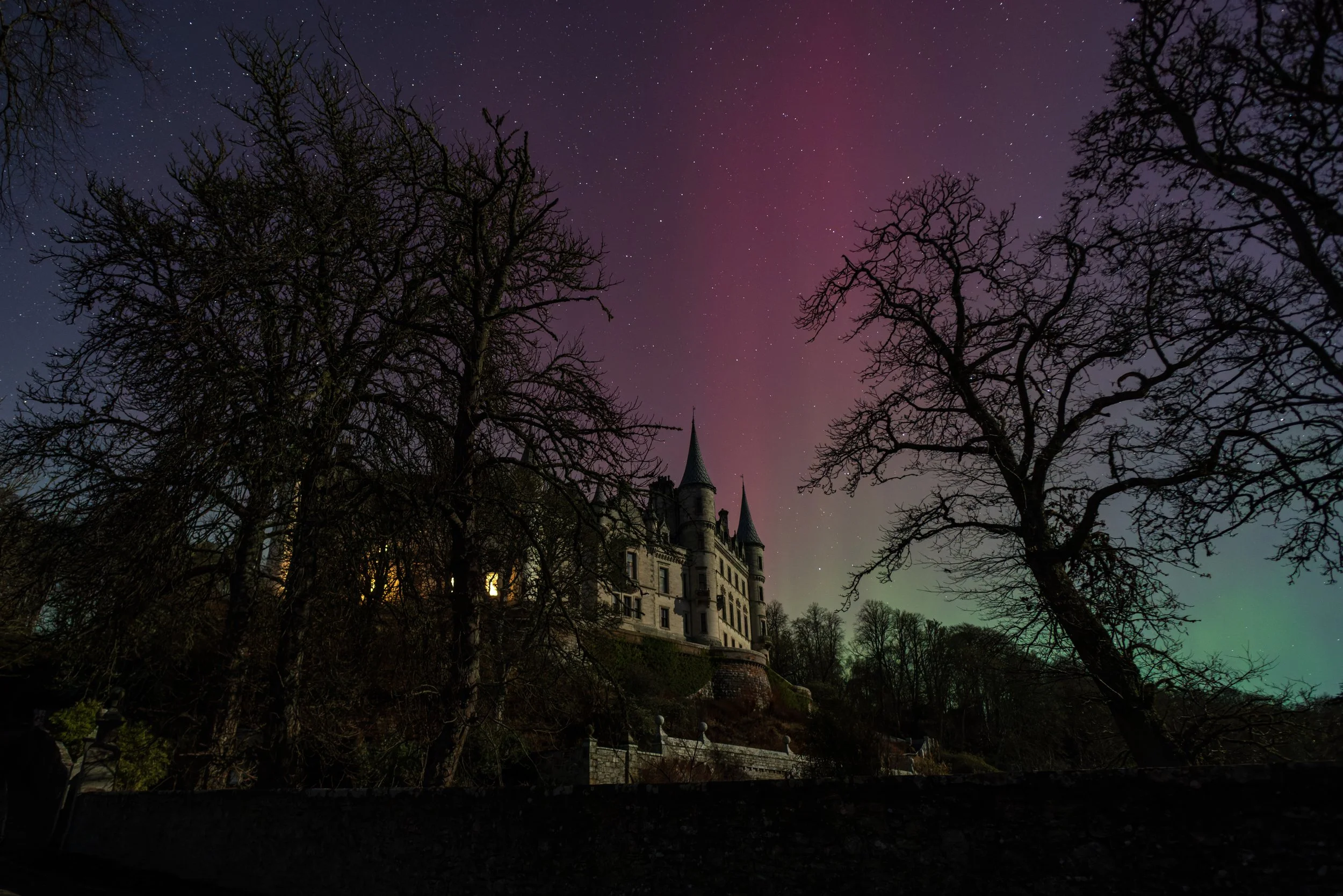 A castle-like building illuminated at night, surrounded by leafless trees, under a starry sky with aurora borealis and a faint purple and green glow.