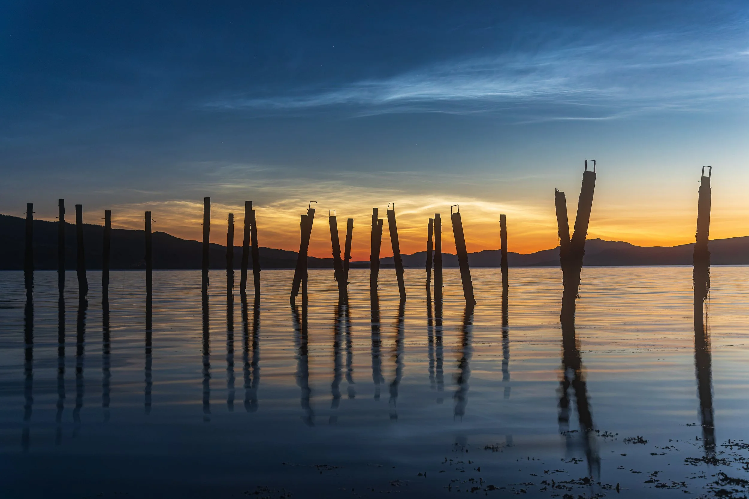 Silhouette of old wooden poles in the water at sunset with mountains in the background and reflected in the calm lake.
