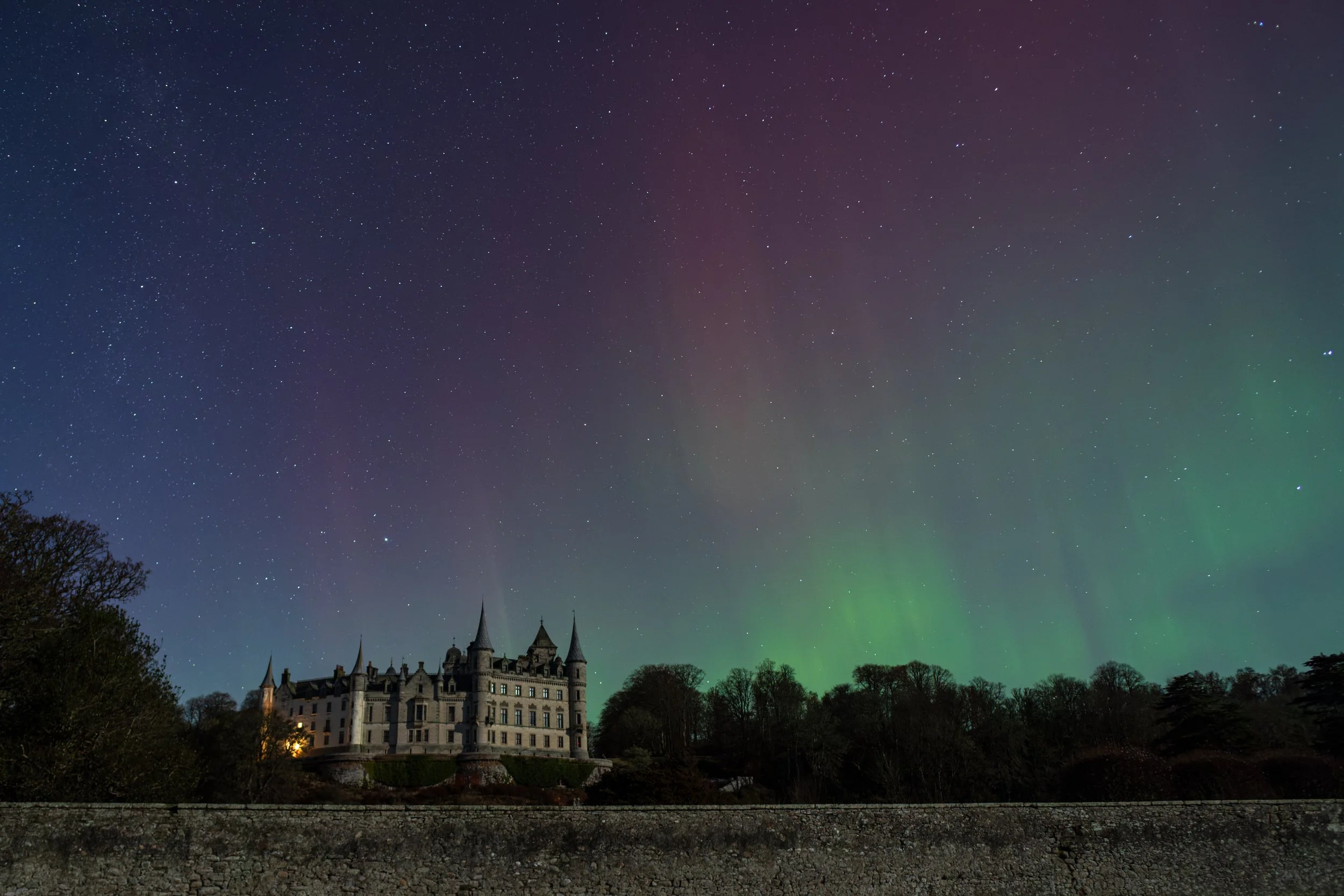 A castle with towers and turrets sits on a hill under a night sky illuminated by the Northern Lights, with trees and a stone wall in the foreground.
