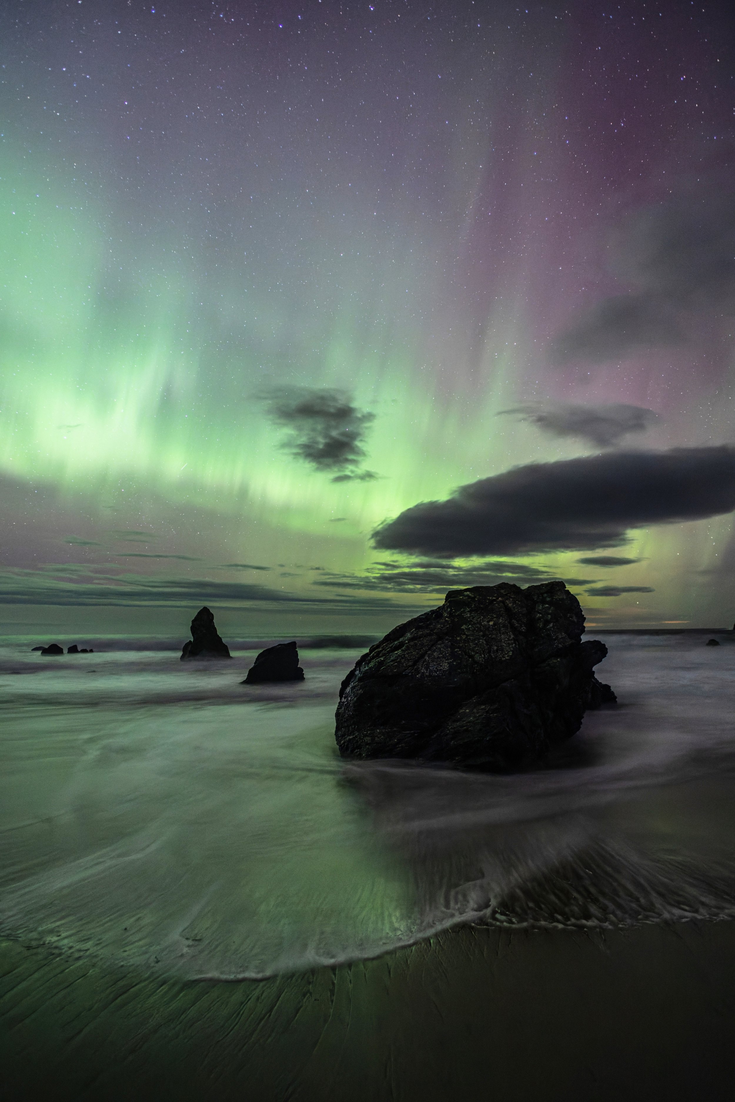 Northern lights over a rocky beach at night with visible starry sky and dark clouds.