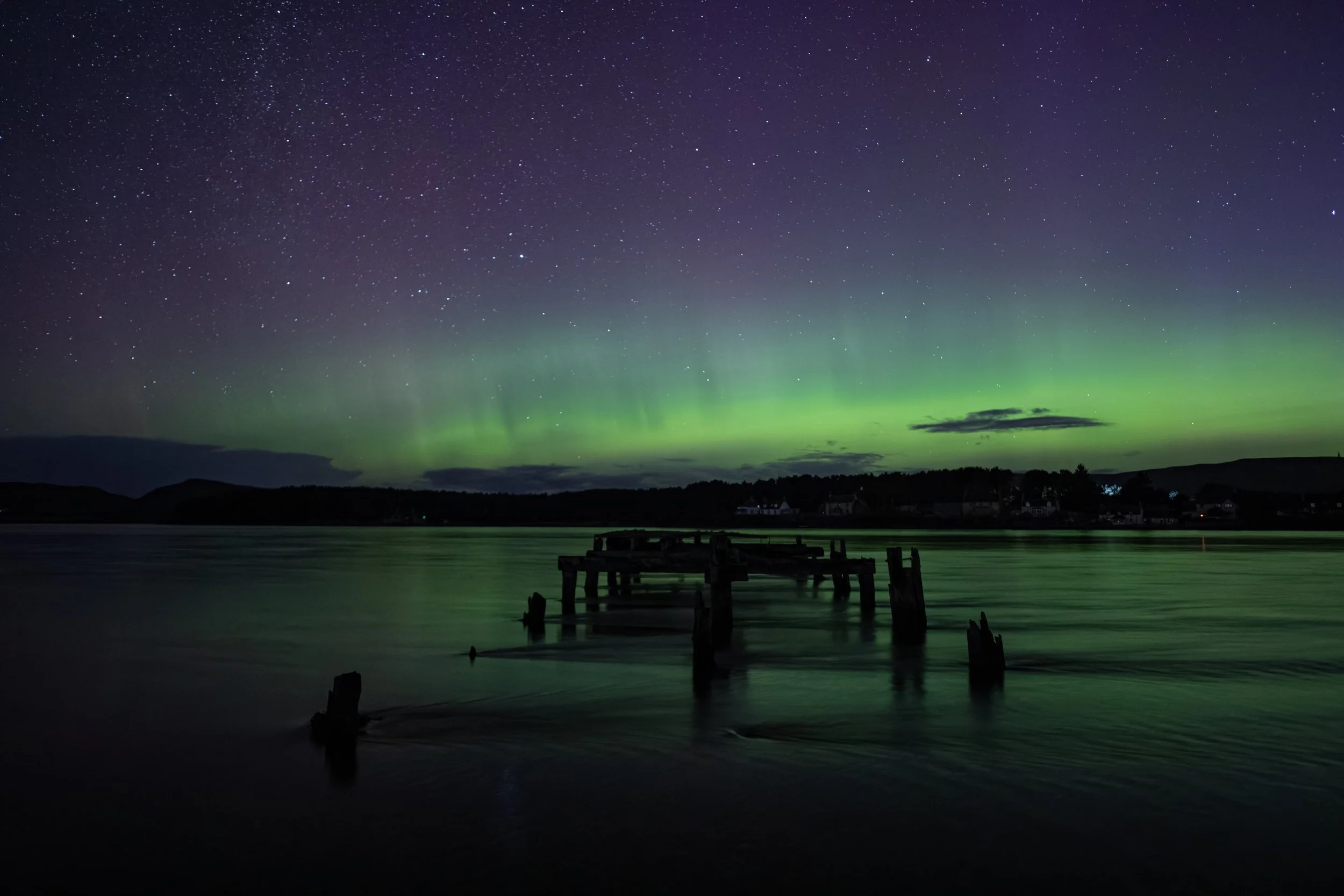 A night scene over a body of water displaying the Northern Lights in green hues across the sky, with stars visible, and remnants of an old wooden pier in the foreground.