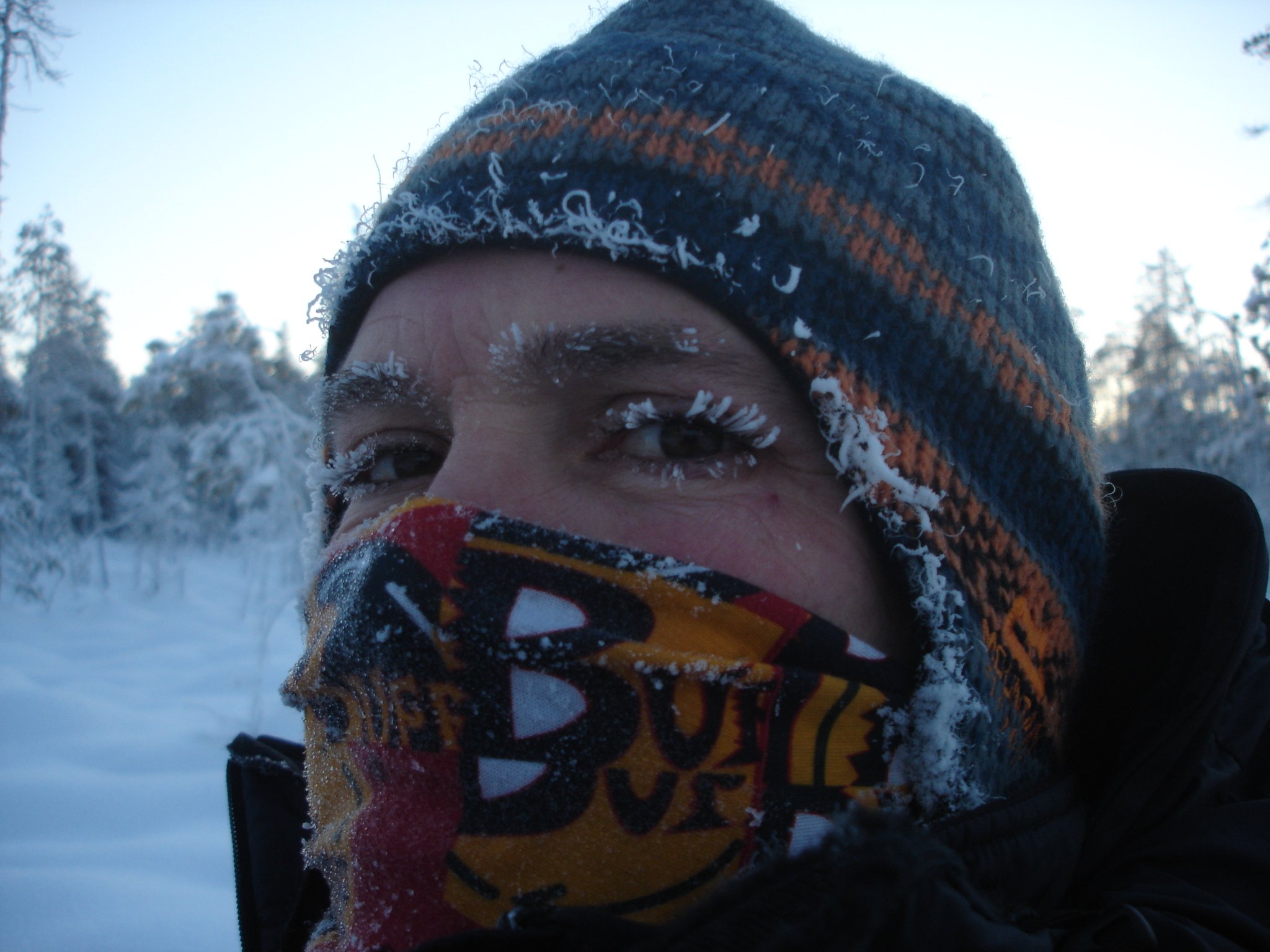 A person wearing a colorful bandana over their face and a blue, gray, and orange striped knit hat in a snowy outdoor setting.
