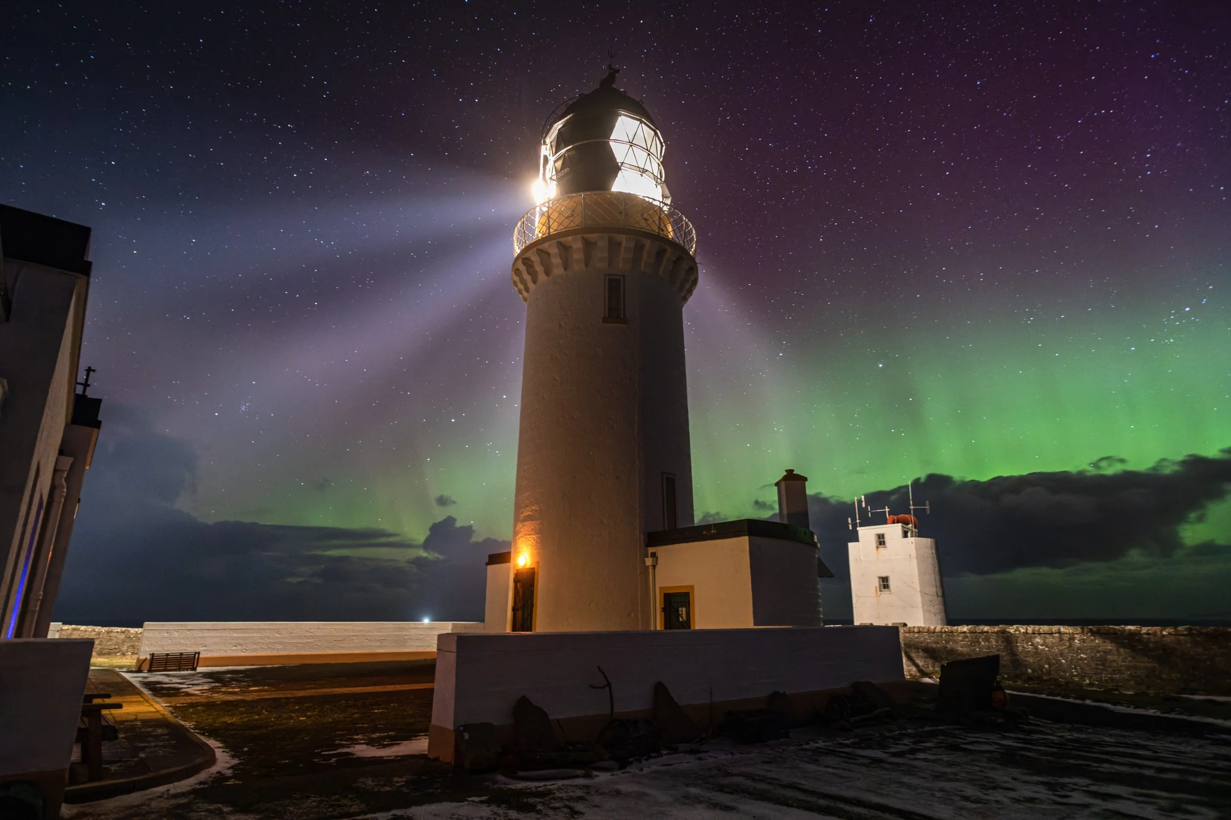 A lighthouse illuminated at night with the Northern Lights and a star-filled sky in the background.