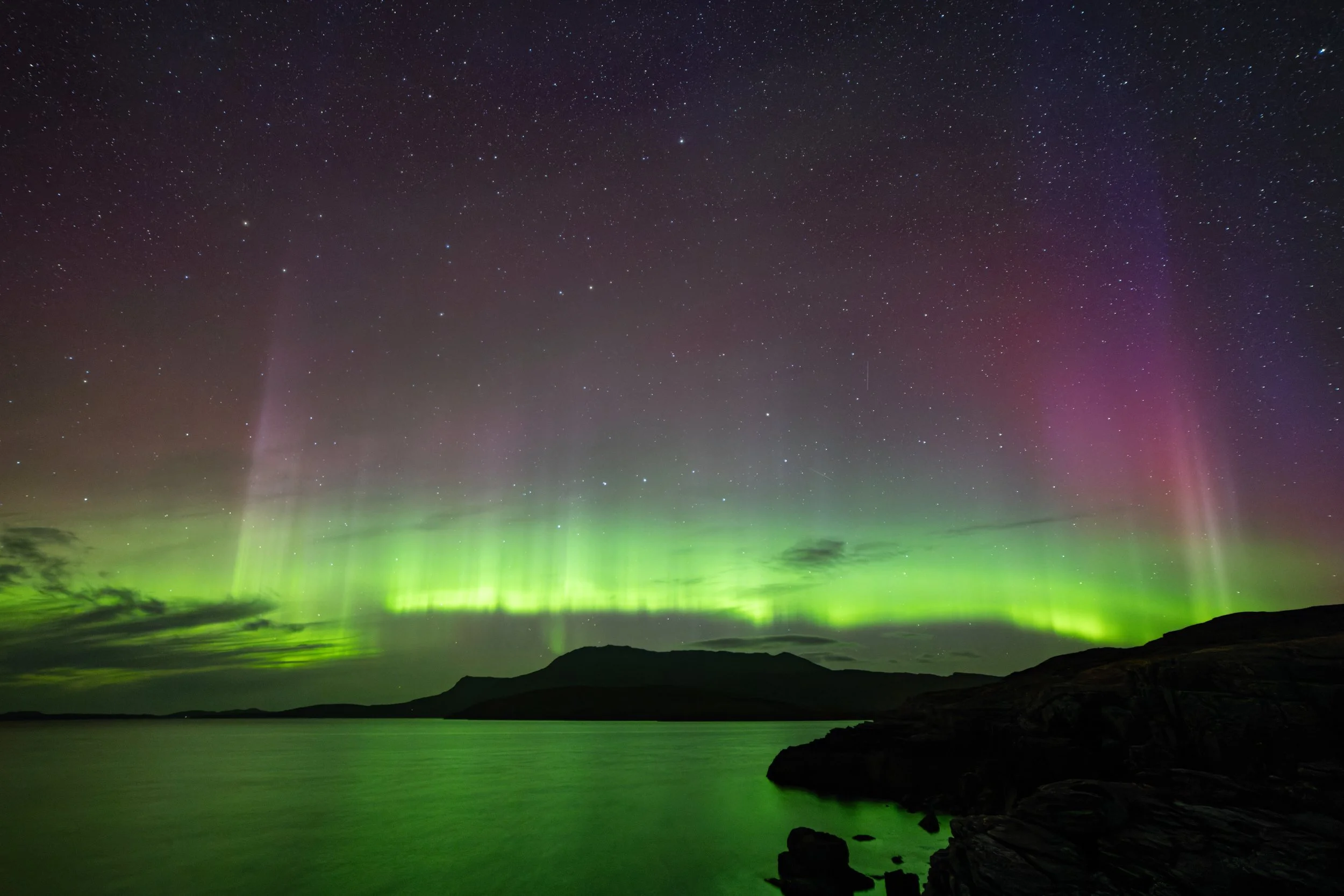 Northern Lights (Aurora Borealis) over a body of water with mountains in the background and a starry night sky.