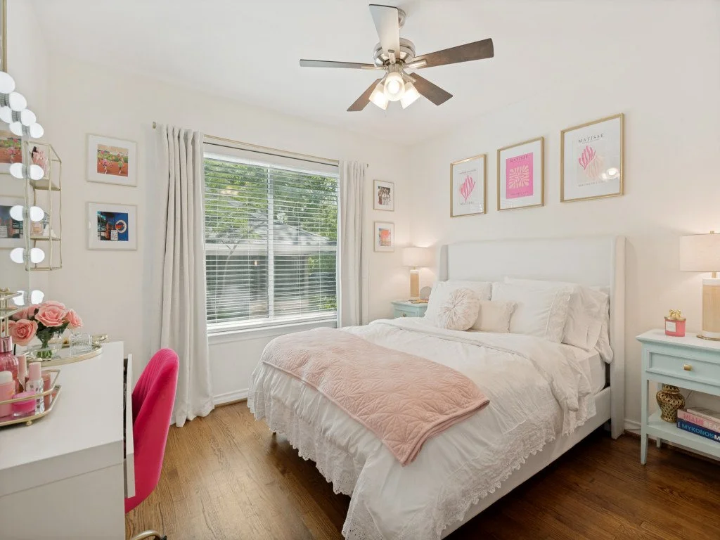 A well-decorated bedroom with a white bed, pink blanket, white pillows, blue nightstands, lamps, and framed artwork on the wall. There is a window with white curtains, a ceiling fan, and a vanity with pink decor on the left.