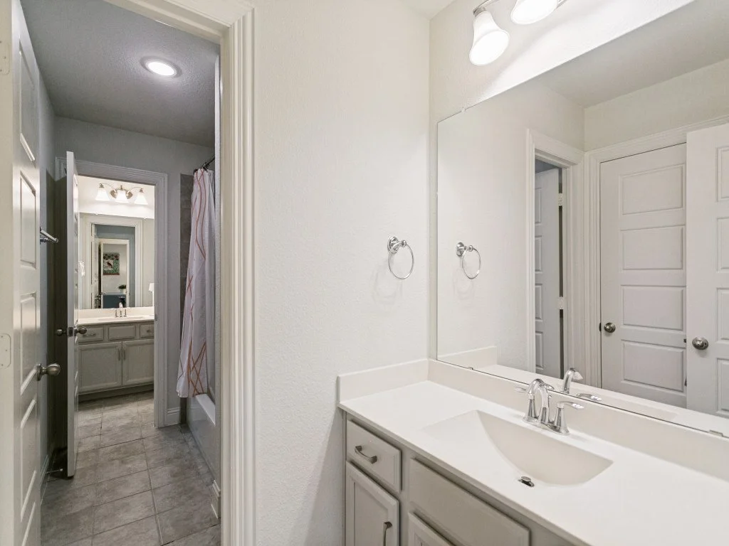 Bathroom with white vanity, sink, and large mirror. Light fixtures above the mirror, beige cabinets, and two towel rings on the wall. Door to a shower or bathtub area with a shower curtain.