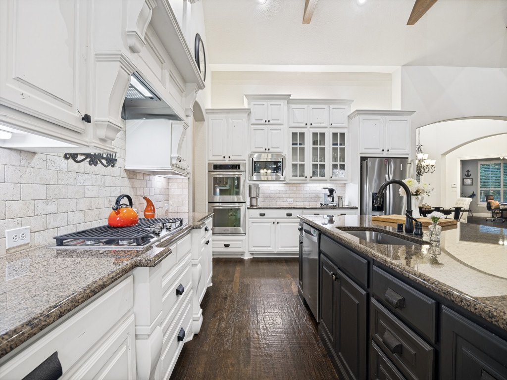 A modern kitchen with white cabinets, granite countertops, and stainless steel appliances including a refrigerator and double oven. The kitchen has a dark island with a sink and a faucet, decorated with white flowers, and an orange kettle on the stov
