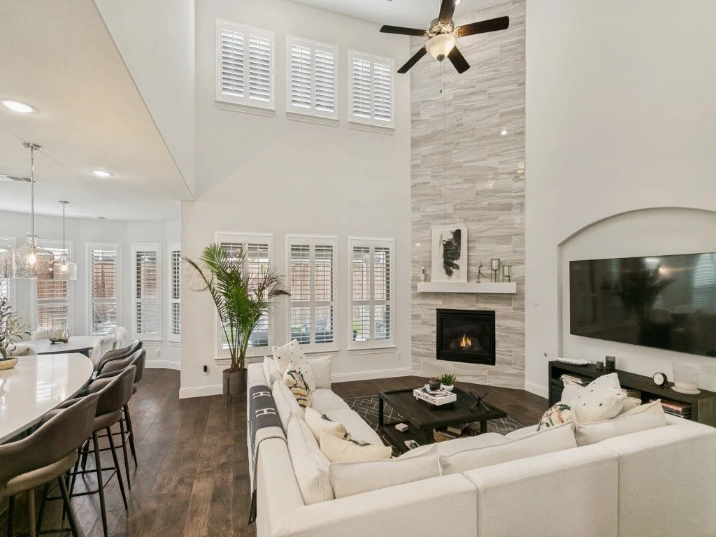 Living room with high ceiling, white walls, a stone accent wall with a fireplace, ceiling fan, large windows with shutters, white sectional sofa, coffee table, and wall-mounted TV.
