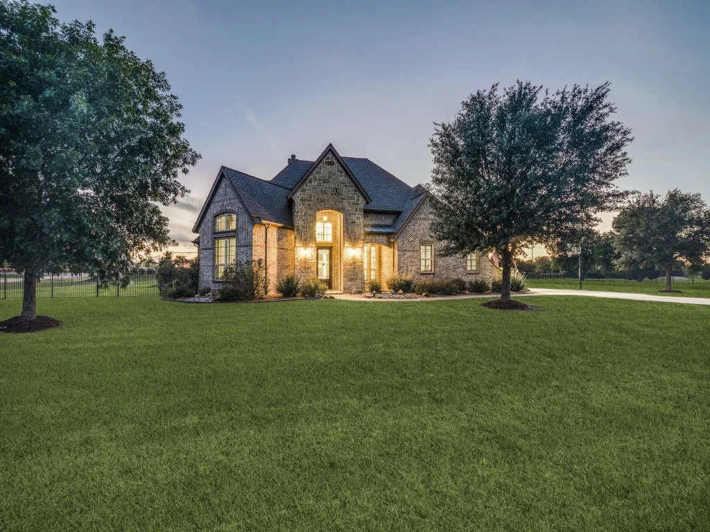 A large brick house with multiple gables, illuminated windows, surrounded by a well-maintained green lawn and trees at dusk.