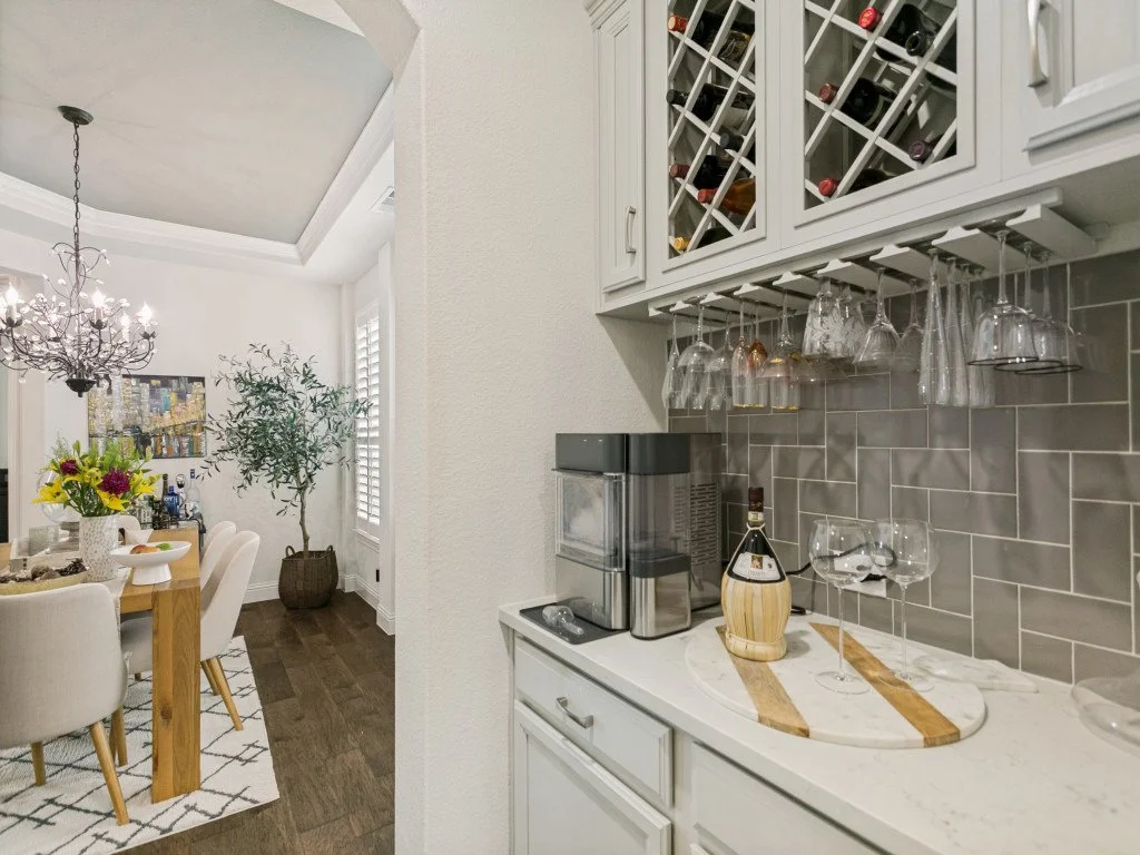 A cozy kitchen nook with hanging wine glasses, a gray tiled backsplash, a small coffee station, and a dining area with a table and cream-colored chairs, floral centerpiece, and a chandelier.