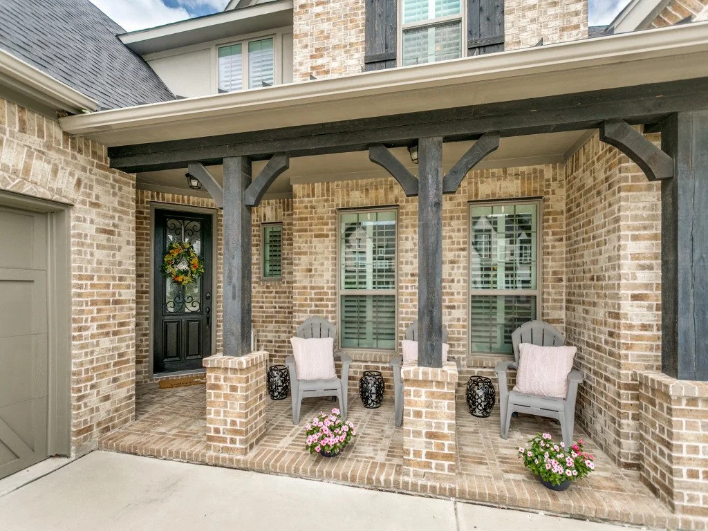 Front porch of a brick house with two chairs, pink pillows, potted flowers, and black lanterns, under a covered area with black wooden support beams.