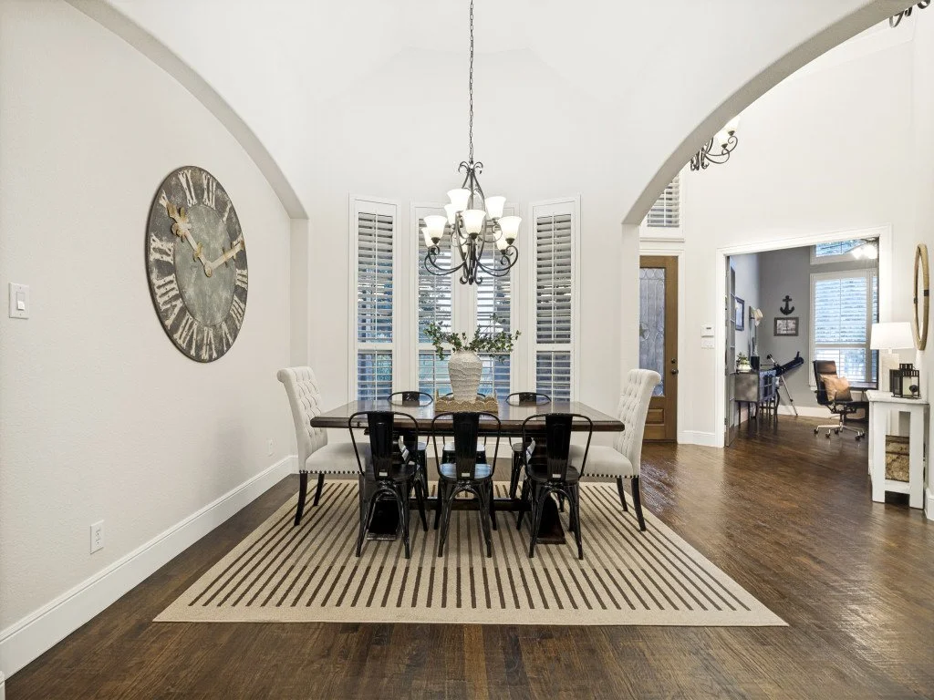 Dining room with a wooden table, six chairs, a chandelier, a large wall clock, and a striped rug. Adjacent to a home office with a desk and chair.