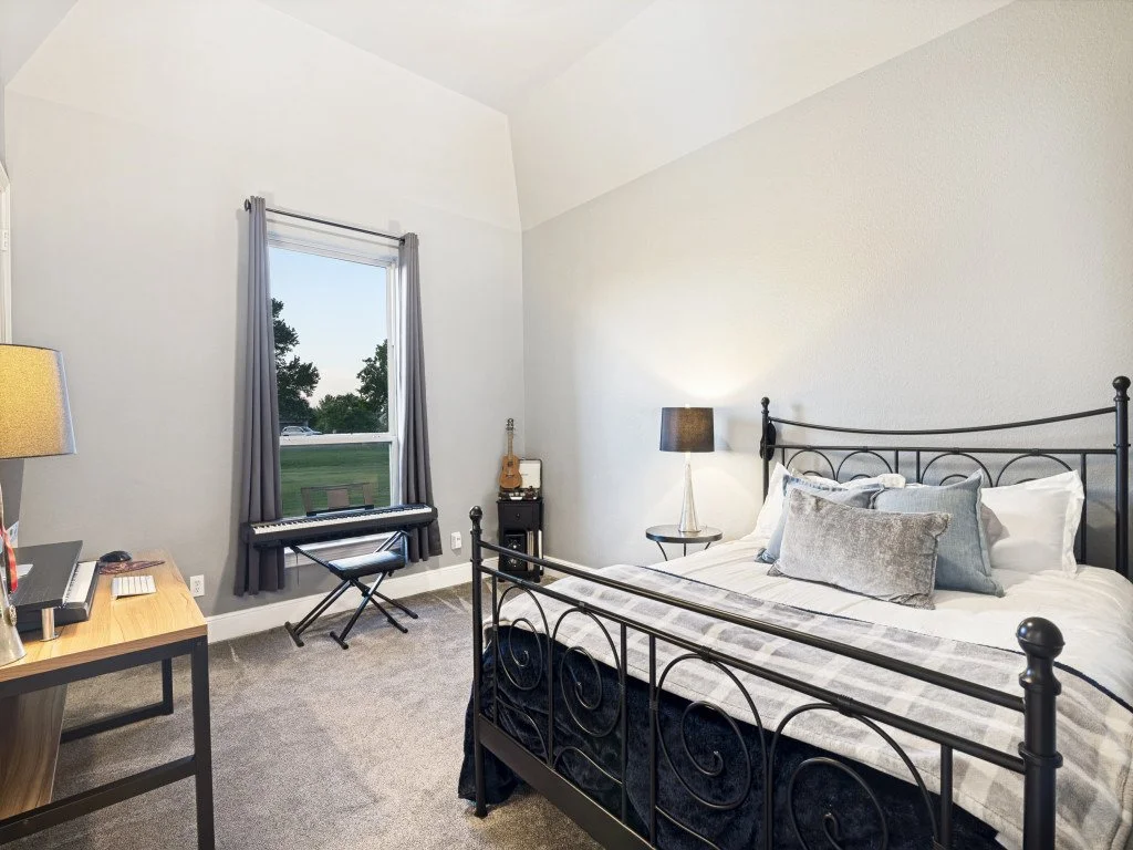 Bedroom with a black metal bed frame, white and gray bedding, nightstand with lamp, window with gray curtains, small keyboard on a stand, and guitar on a small black table.