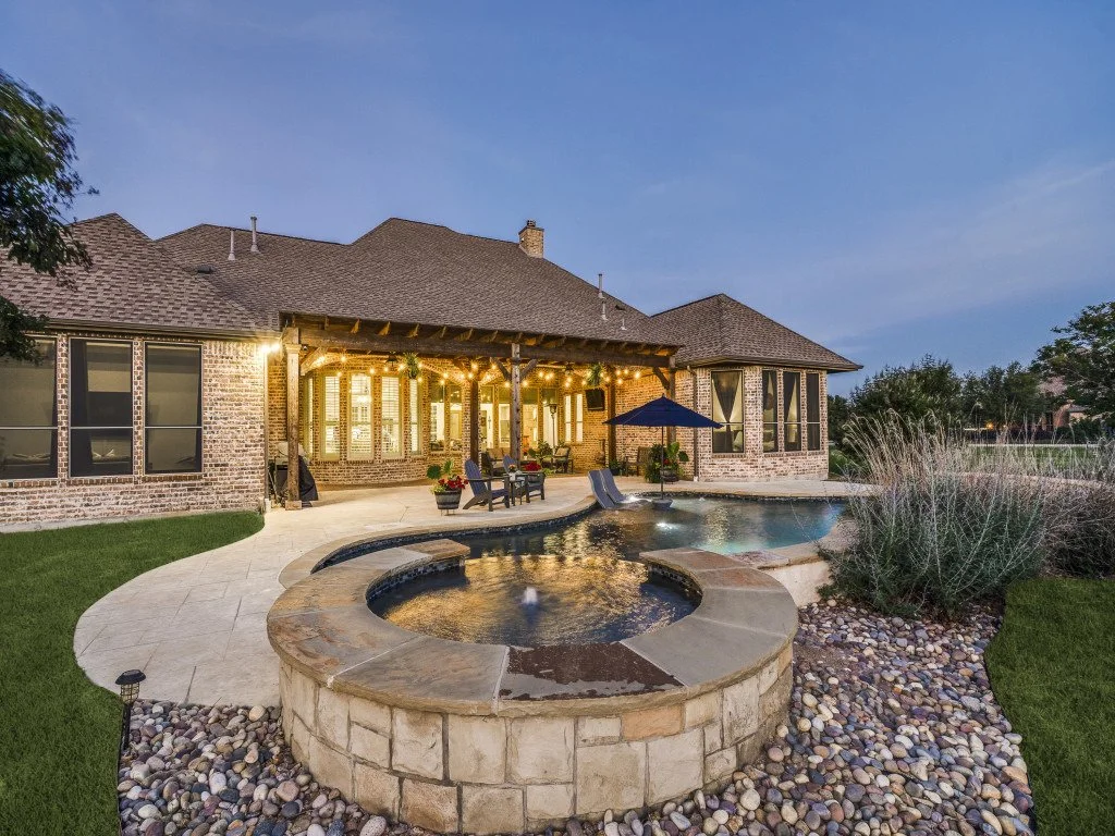 Backyard with a lit patio, swimming pool, hot tub, outdoor furniture, and landscaped yard at dusk.