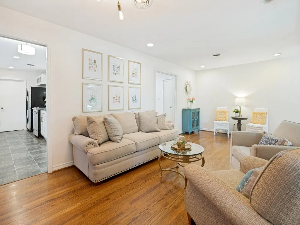 Living room with beige sofa, armchair, coffee table, two chairs with yellow pillows, blue cabinet, side table with lamp, wall art, hardwood floor, and doorway leading to kitchen.