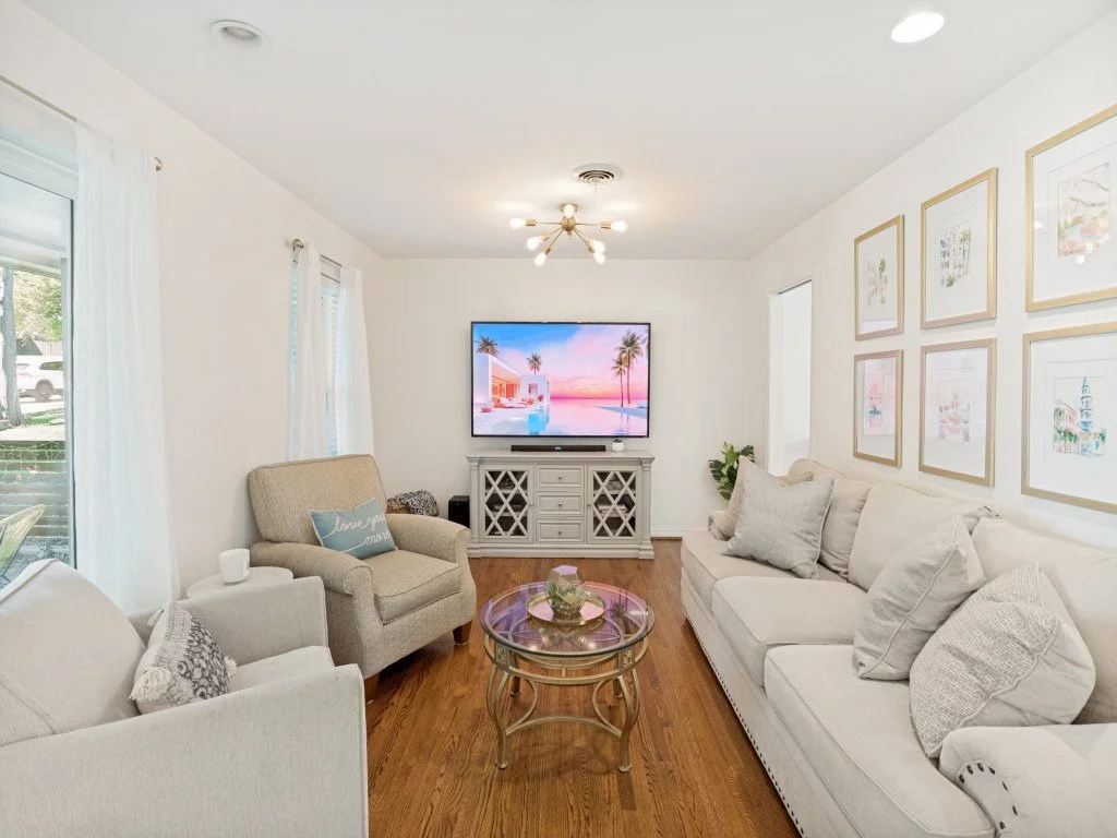 Living room with white walls and wooden floor, beige couches, a glass coffee table, a large flat-screen TV on a white TV stand, framed artwork on the wall, and windows with white curtains.