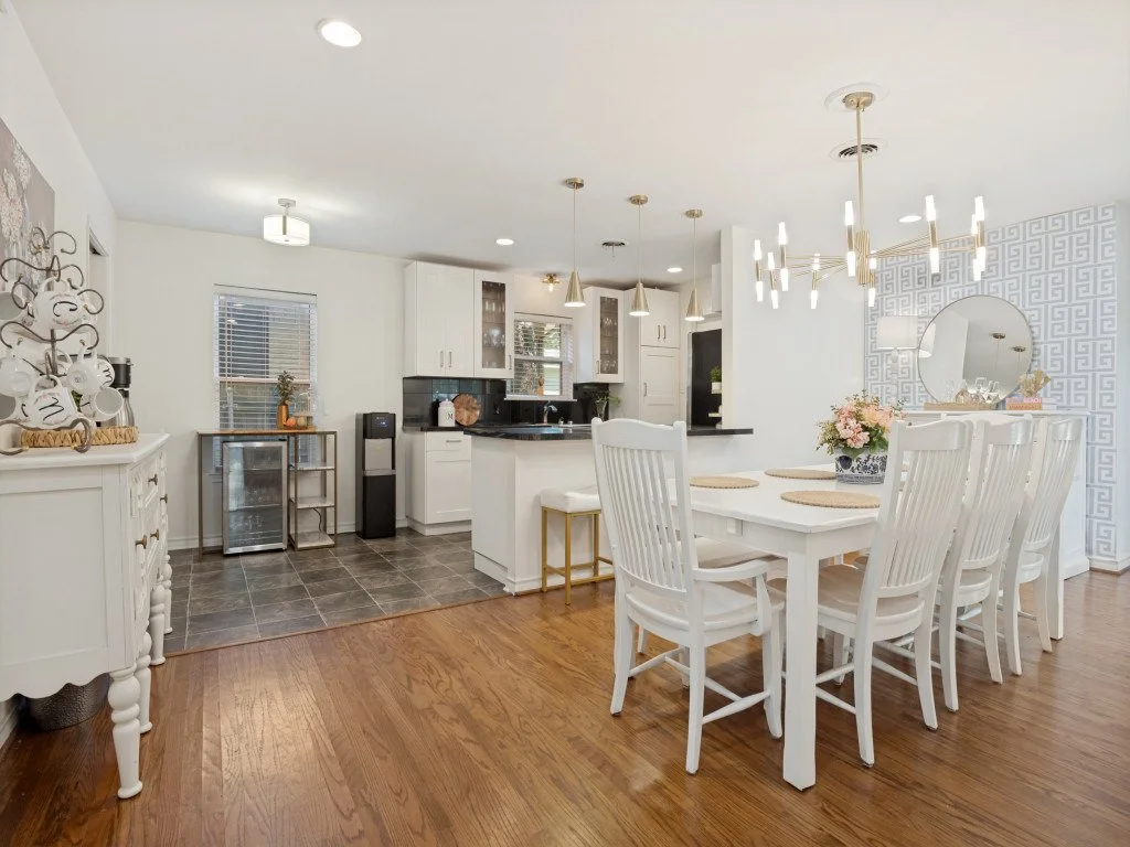 Open-plan kitchen and dining area with white furniture, black countertops, hardwood floor, and modern light fixtures.