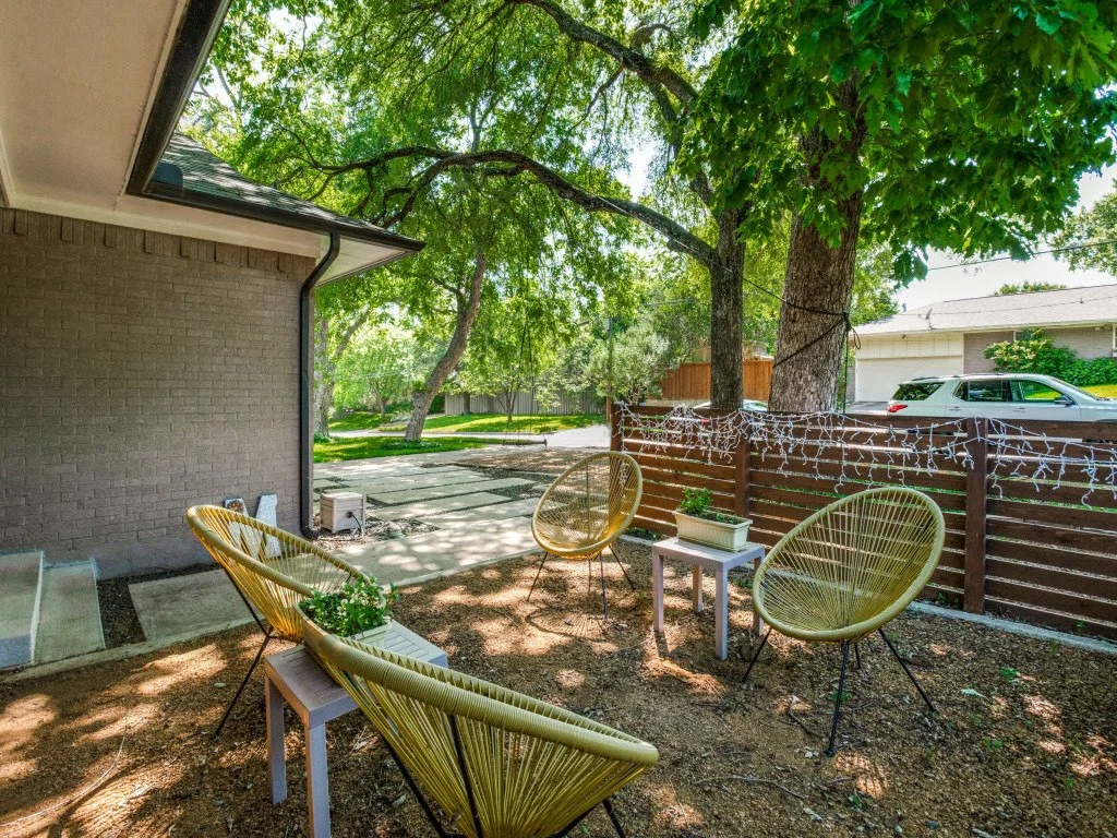 A backyard patio with four yellow circular chairs around a small white table, a large tree providing shade, and string lights hanging along a brown wooden fence. There are green plants in white planters on the tables, and the scene is partly shaded w