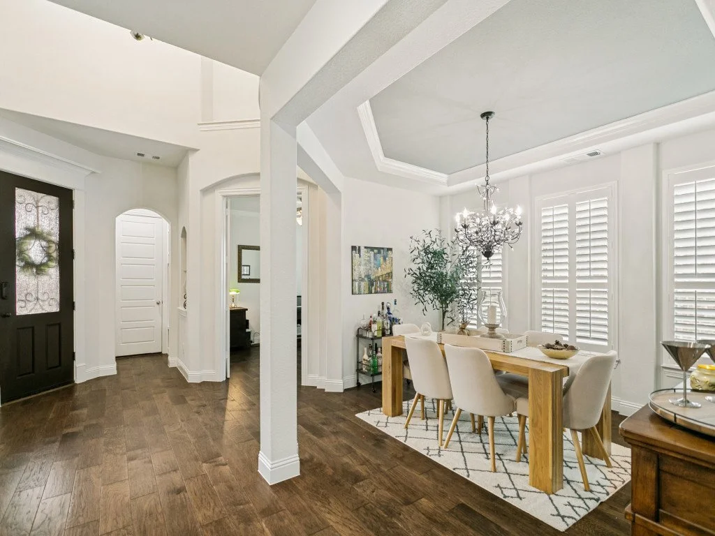 A dining area with a wooden table, six white upholstered chairs, a chandelier hanging from the ceiling, a rug underneath the table, and large windows with white shutters. There is a side table with a tray and decorative items. The room has white wall