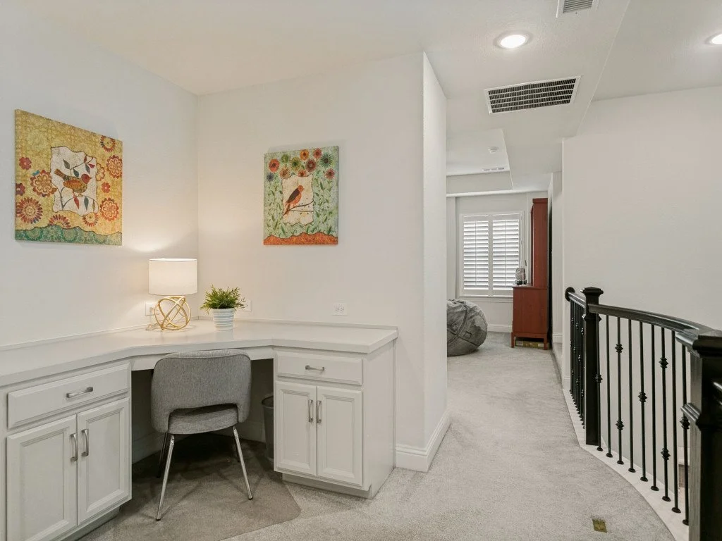 A bright, modern interior with a built-in white desk and gray chair. Decor includes wall art with colorful nature scenes, a table lamp, and a small plant. Visible hallway with black railing and a room with window shutters and a cabinet in the backgro