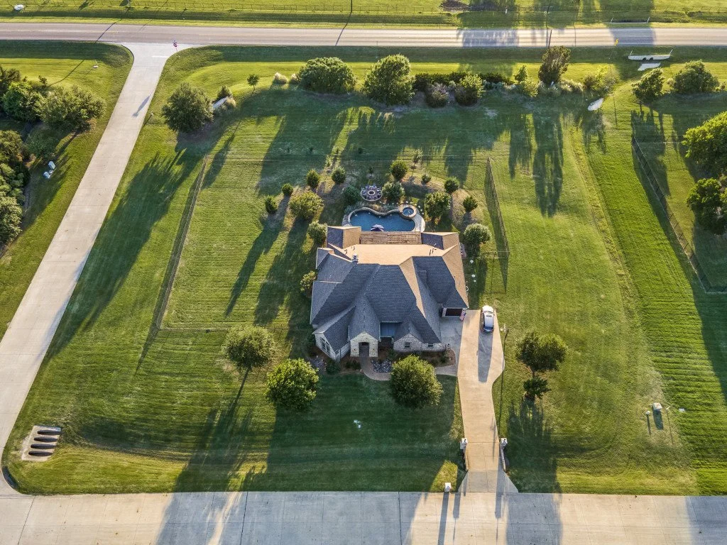 Aerial view of a large house with a curved driveway, surrounded by green lawns with trees, a swimming pool, and a fenced backyard. Street roads border the property on two sides.