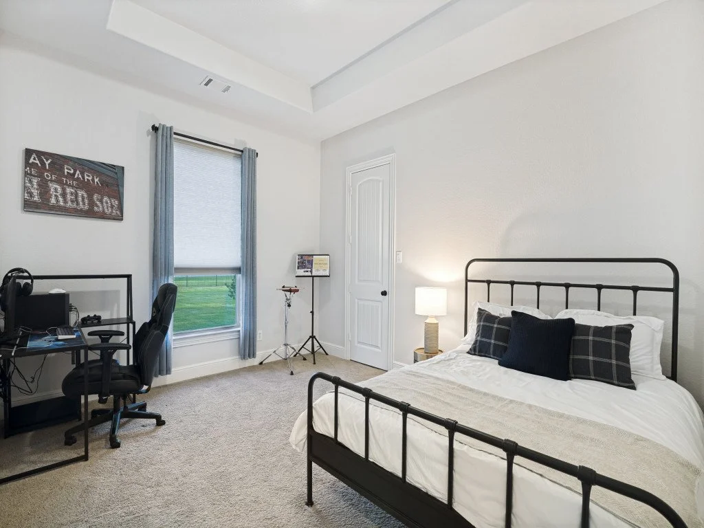Bedroom with a black metal bed frame, three pillows, a nightstand with a lamp, a window with blue curtains, a computer desk with an office chair, and a sign on the wall.