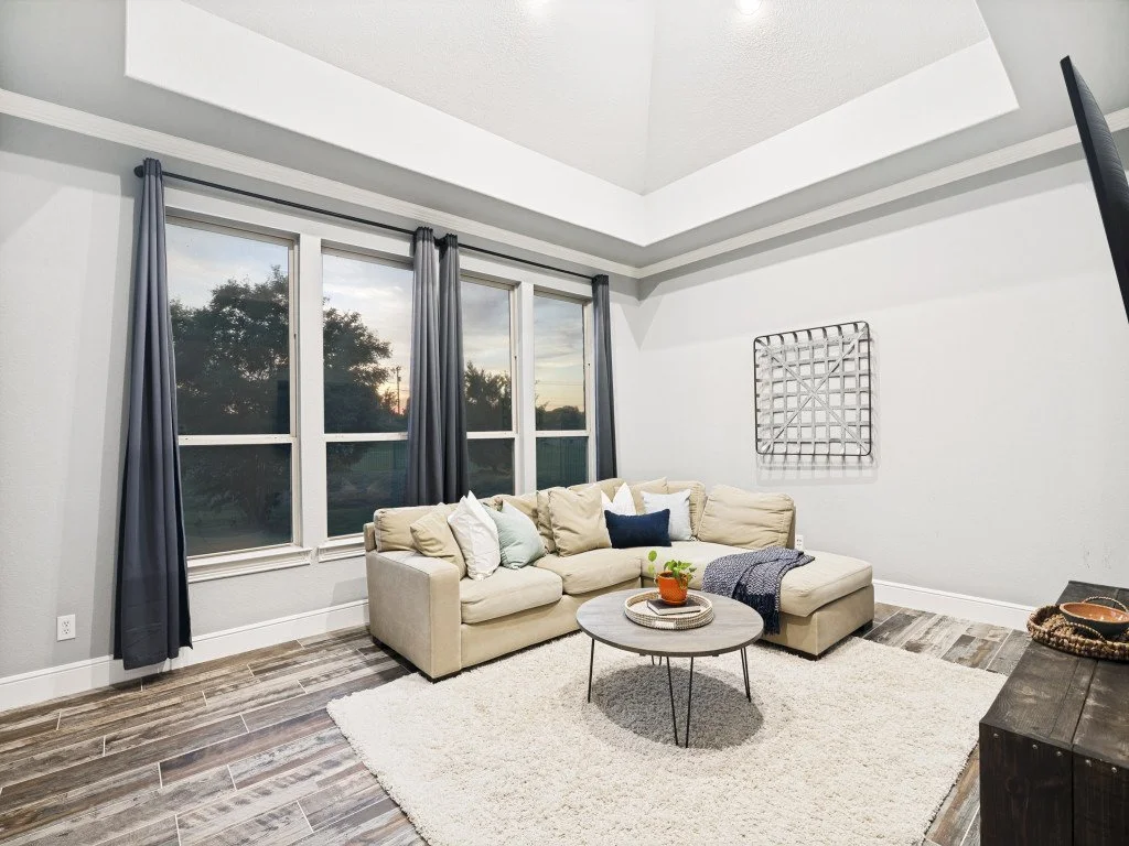 Living room with large windows, beige sectional sofa with pillows, round coffee table, dark wood side table, and a white textured rug.