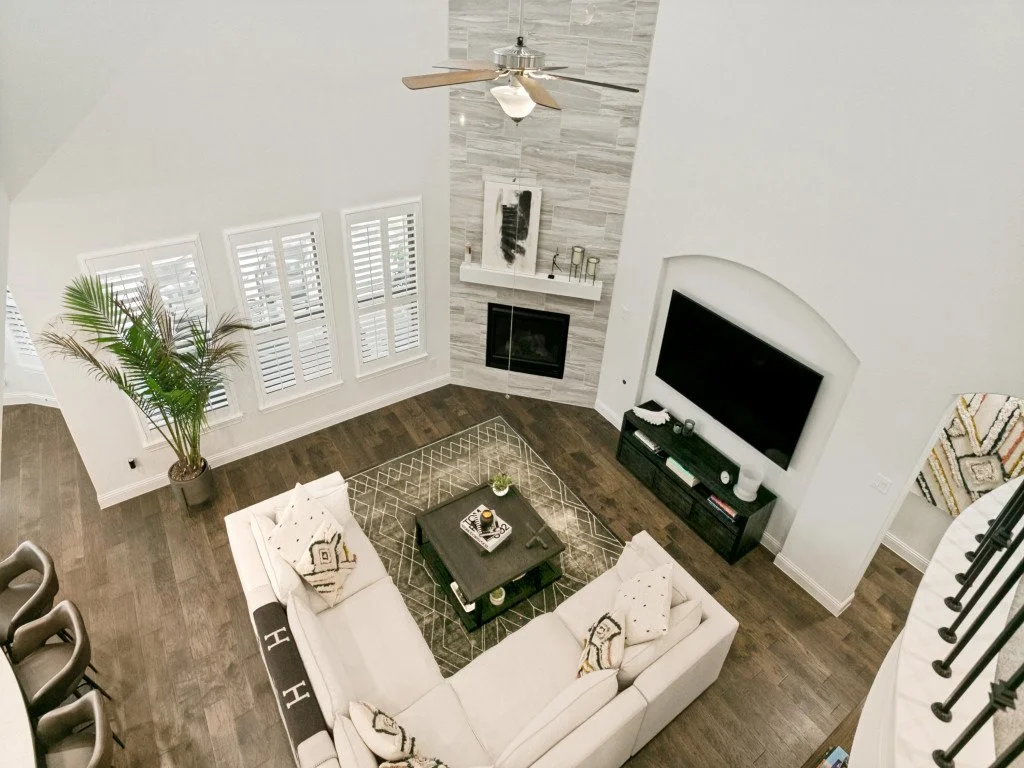 Living room with a white sectional sofa, a black coffee table, a gray area rug, a fireplace with a stone surround, a large flat-screen TV, a tall potted plant, and a ceiling fan.