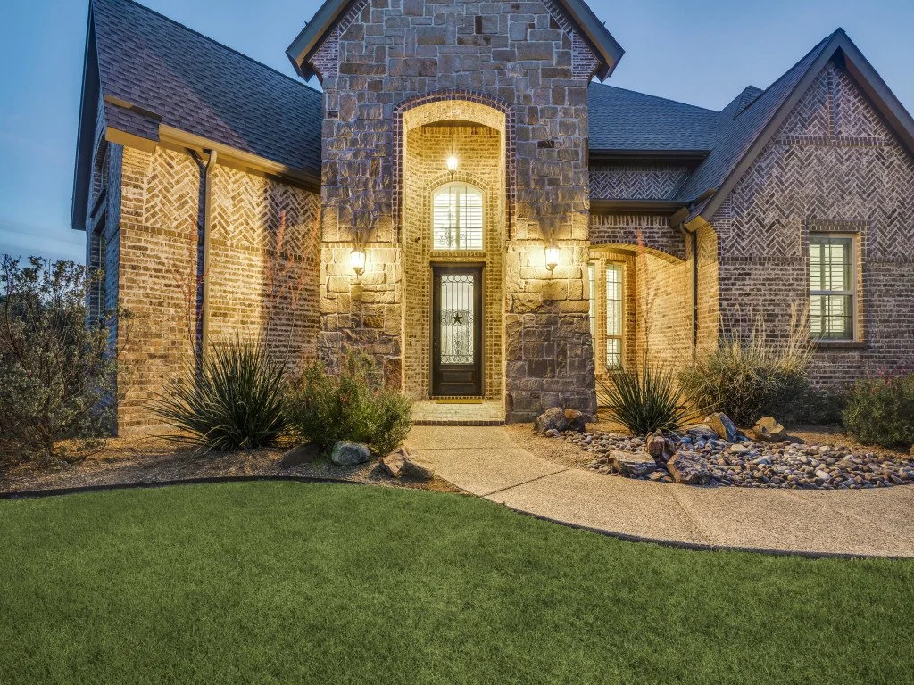 Front view of a brick house with stone accents, illuminated by exterior lights during dusk.