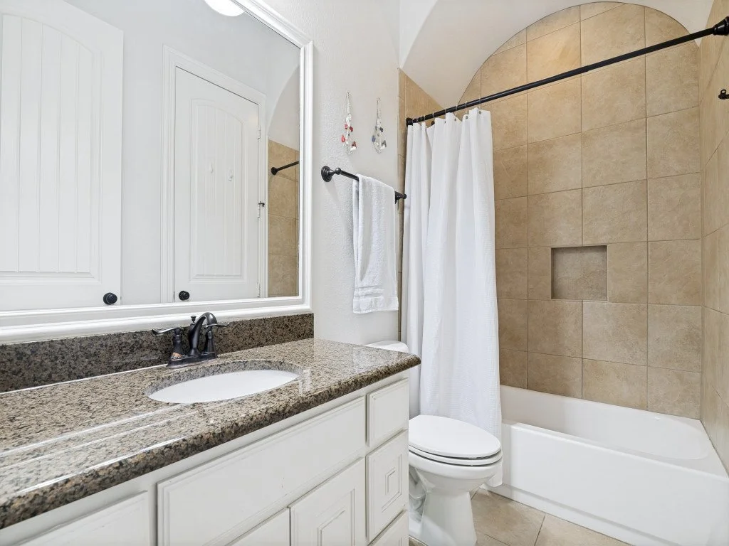 Bathroom with beige-tiled shower, white bathtub, white vanity with granite countertop, mirror, and white towel