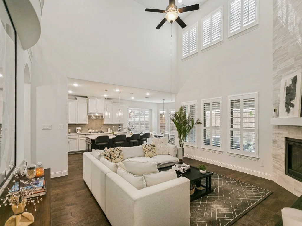 Open-concept living room and kitchen with white walls, high ceiling, large windows, ceiling fan, white sectional sofa, black coffee table, fireplace with light gray stone, dark wood flooring, and bar with black stools.